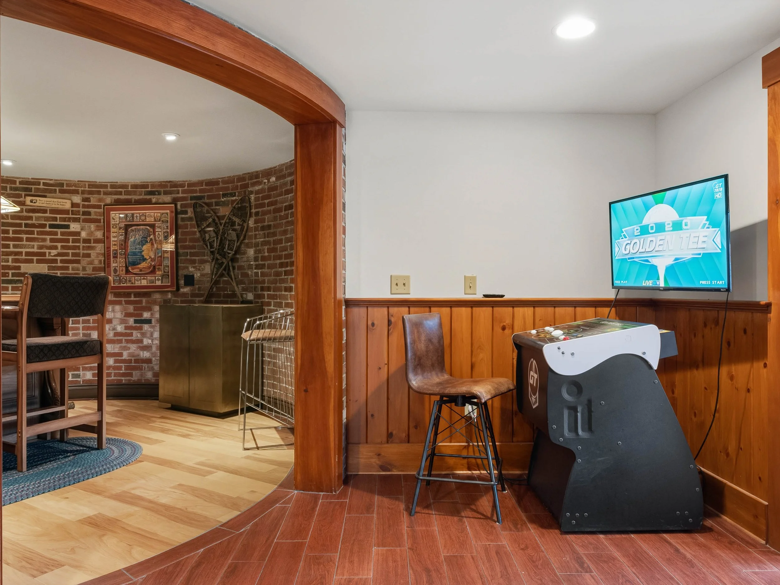 Home arcade setup with a stool, arcade machine, and TV on a wooden-paneled wall in a room with red wood flooring.