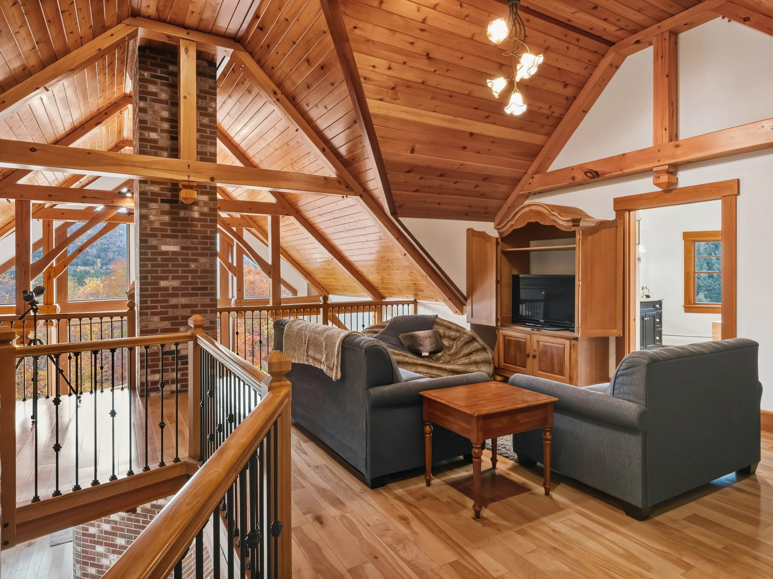 Cozy living room in a log cabin with wood-paneled ceiling, brick chimney, and large windows showcasing fall foliage, furnished with gray sofas, a wooden coffee table, and a wooden entertainment center.