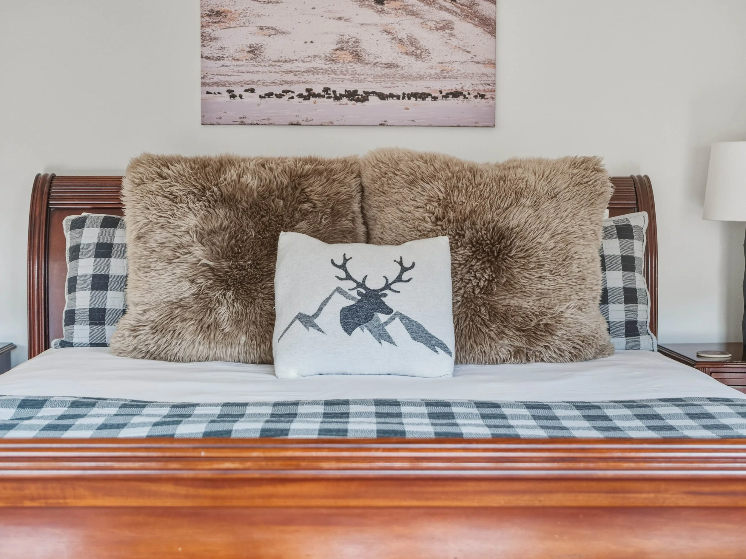 A bedroom with a wooden bedframe, topped with a white sheet, plaid pillows, a large furry pillow, and a decorative pillow with a mountain and deer head design. There is a landscape painting on the wall above the bed and a lamp on the nightstand to th