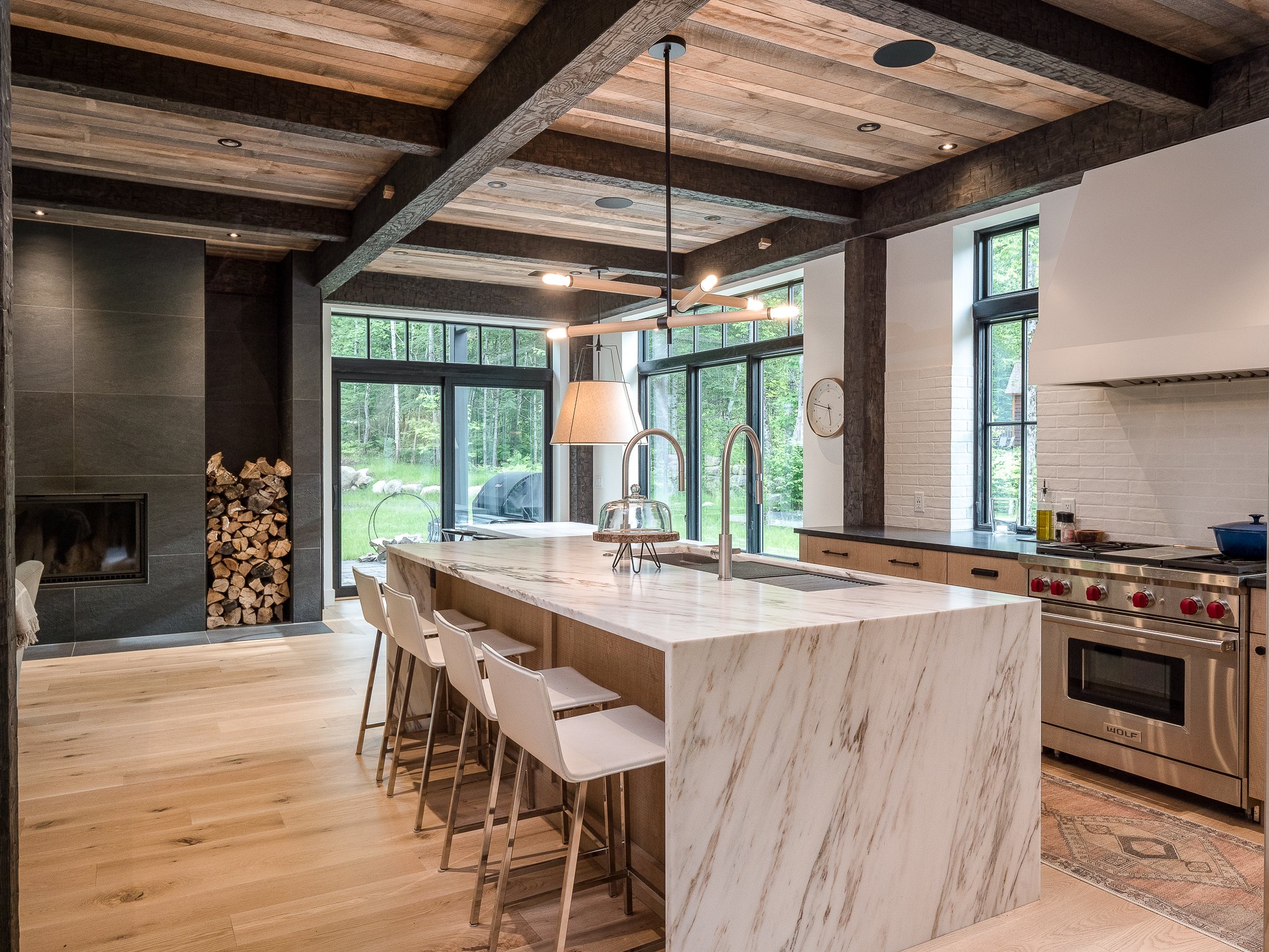 Modern kitchen with marble island, wooden beams, bar stools, large windows, fireplace with firewood, and stainless steel appliances.