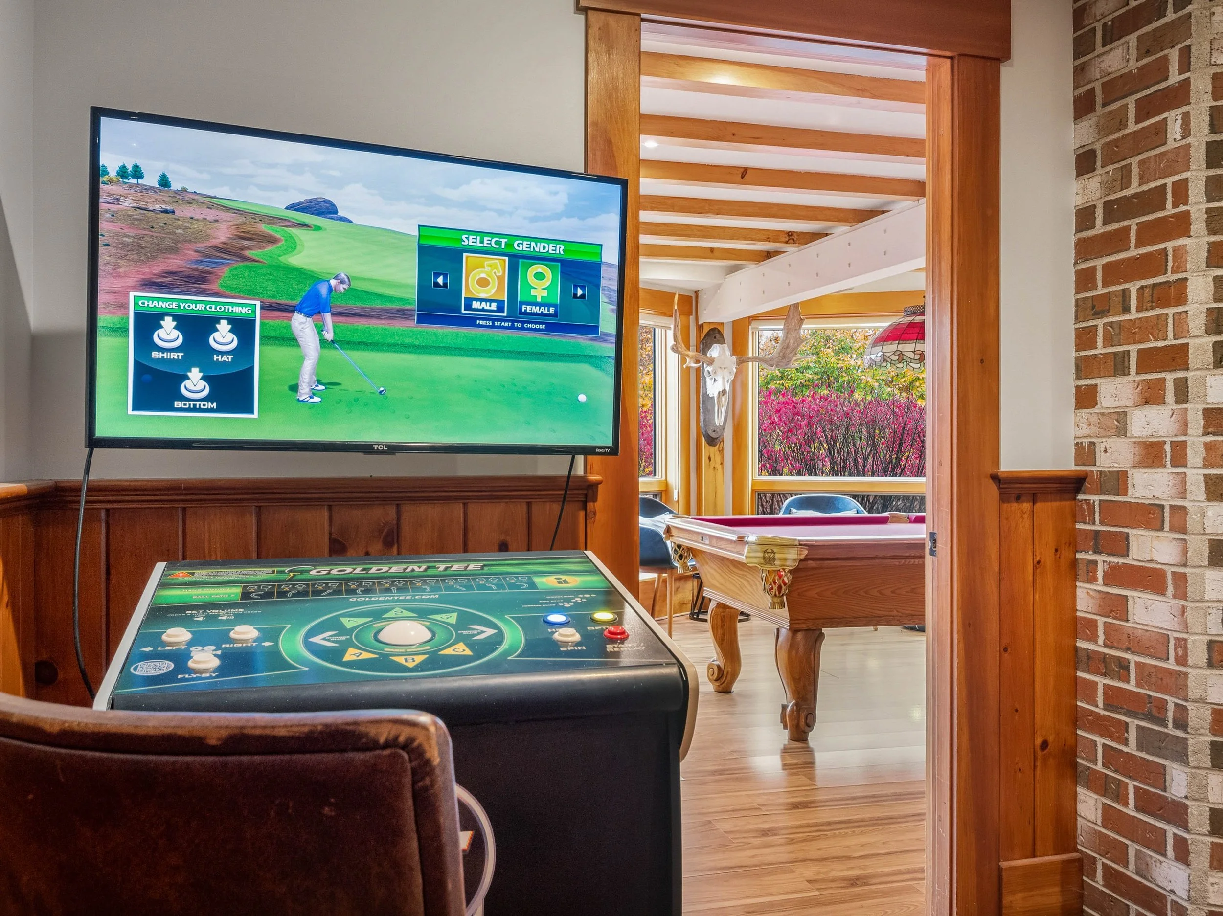 Indoor recreational room with a television showing a golf video game, an arcade-style golf game controller, a pool table in the background, and a window with pink flowering trees outside.