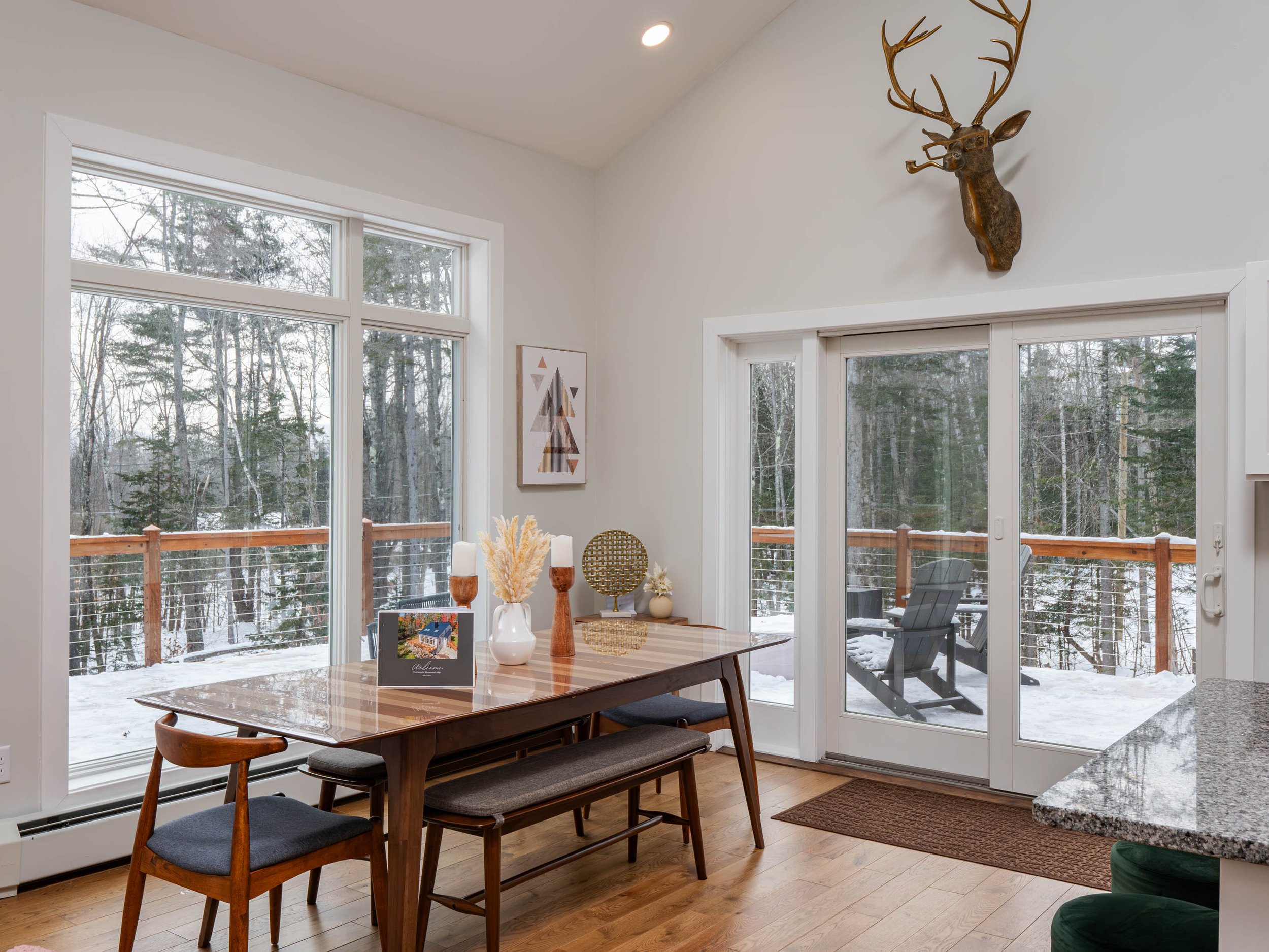 Dining area with large windows and glass sliding door overlooking a snowy wooded outdoor deck, decorated with a mounted deer head on the wall.