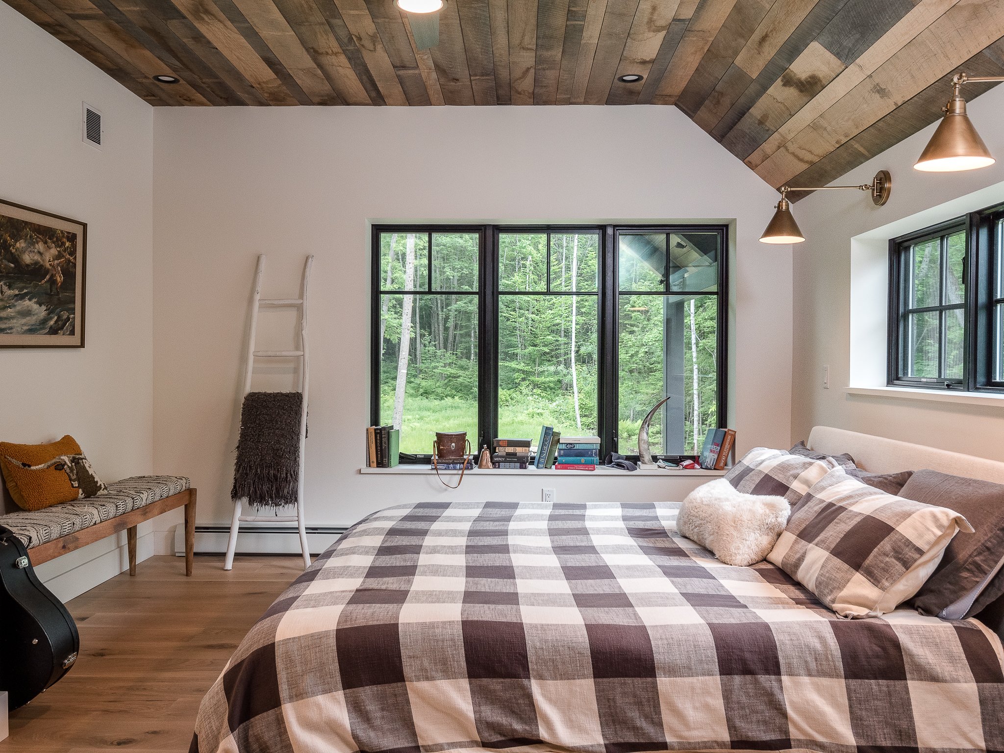 Cozy bedroom with checkered bedspread, wooden ceiling, large window view of forest, bench with cushions, decorative ladder, and books on windowsill.