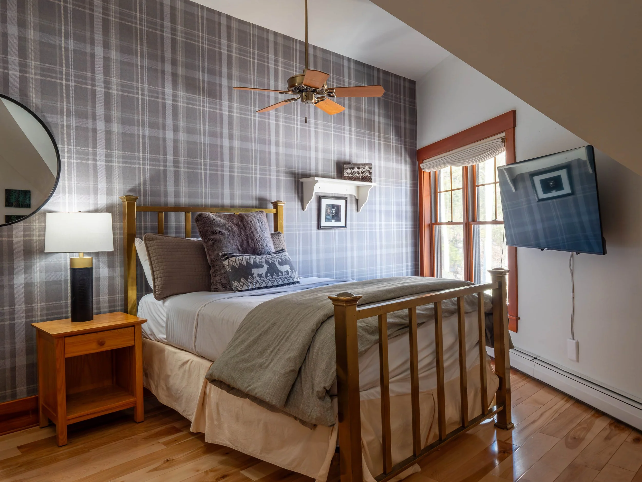 A cozy bedroom with a bed, wooden nightstand, wall-mounted TV, plaid wallpaper, window with wooden trim, ceiling fan, and soft decor.