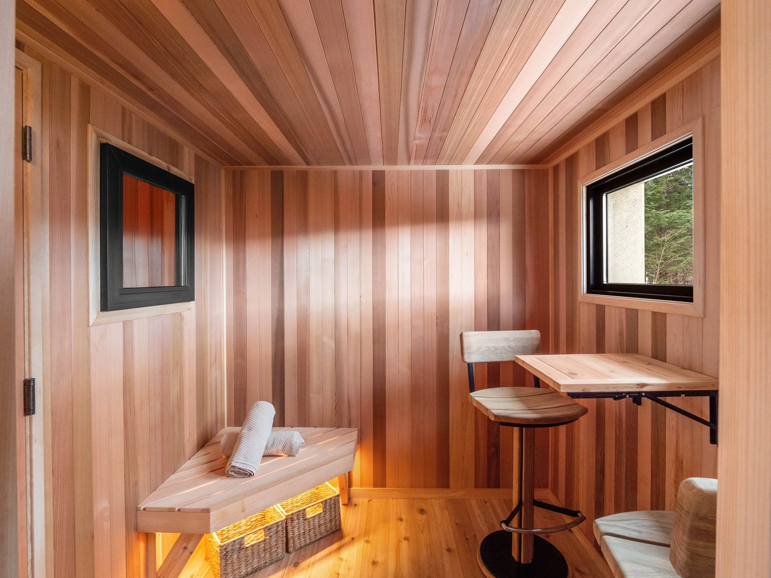 Small wooden room with natural wood-paneled walls, ceiling, and floor. There are two windows with black frames, one on each wall. The room contains a small wooden table with a matching chair and a basket underneath. A towel is neatly rolled on a wood