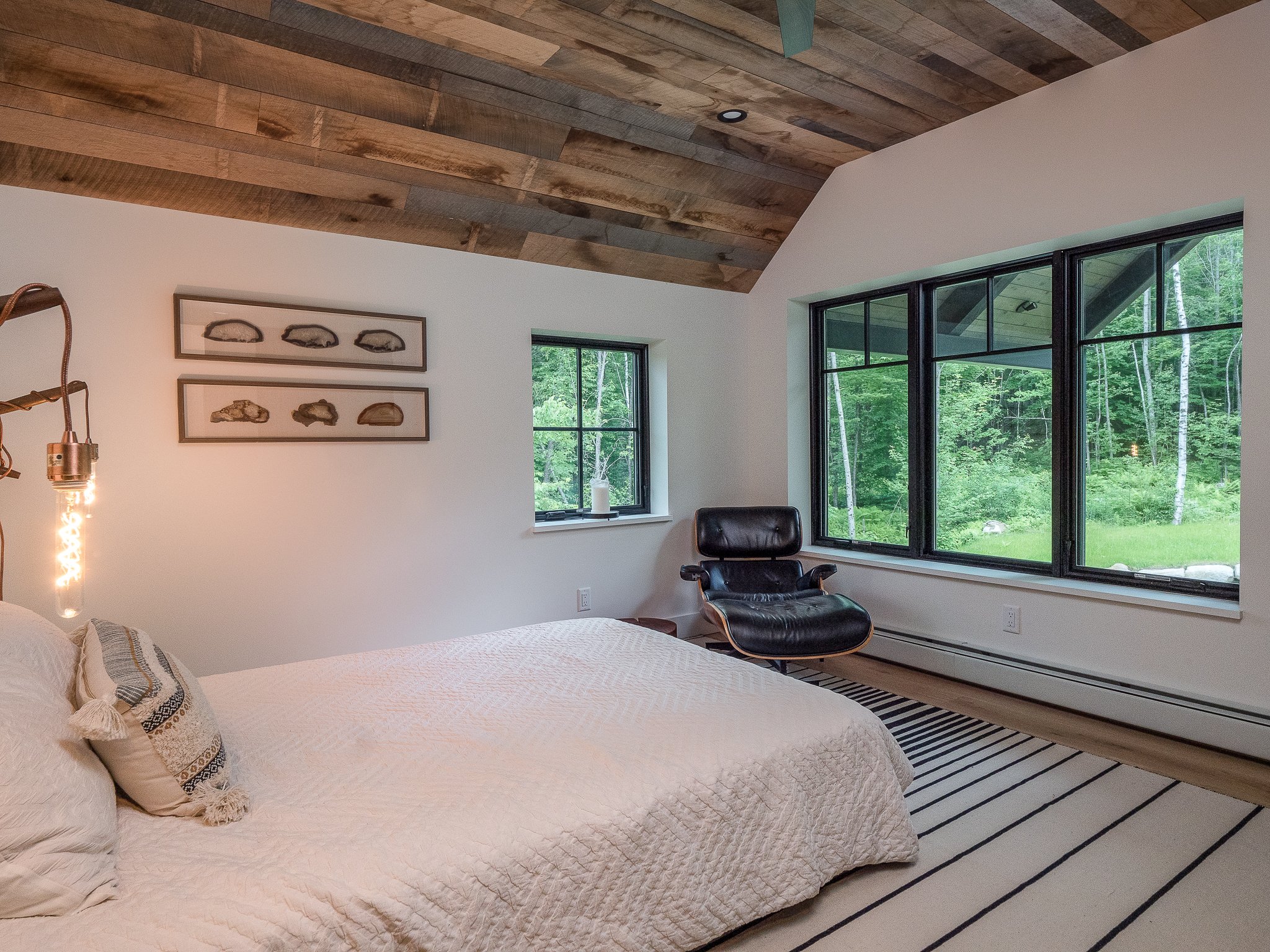 Modern bedroom with wooden ceiling, white bedspread, decorative wall art, large windows, black lounge chair, and striped rug.