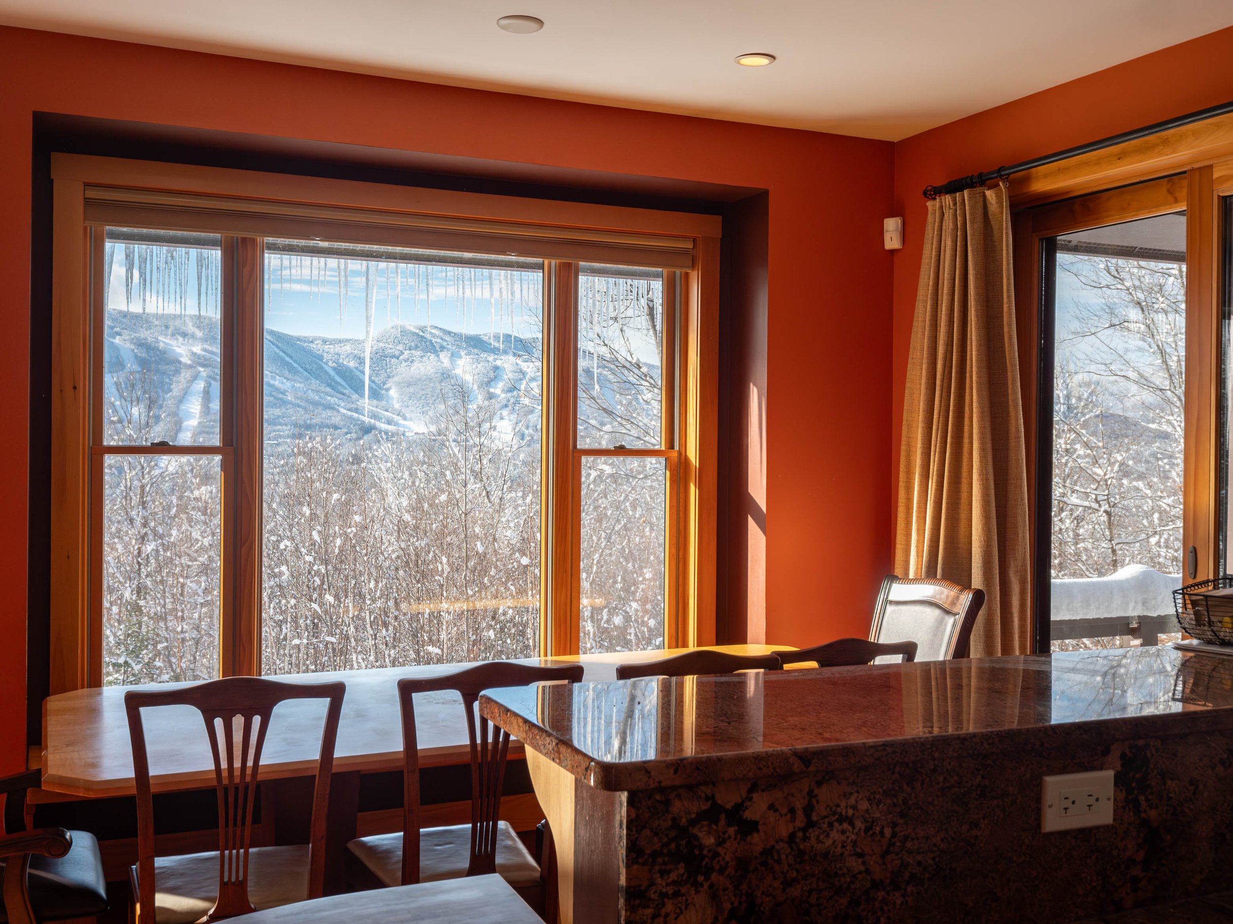Interior view of a dining area with a large window showing a snowy mountain landscape. The room has orange walls, a wooden table and chairs, granite countertop, and curtains.