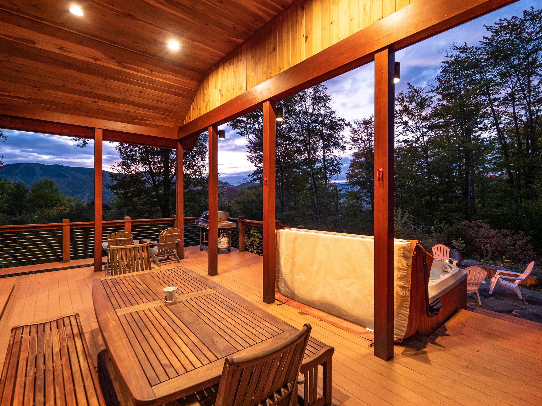 Outdoor wooden deck with hot tub, wooden table, chairs, patio furniture, view of trees and mountains at sunset.
