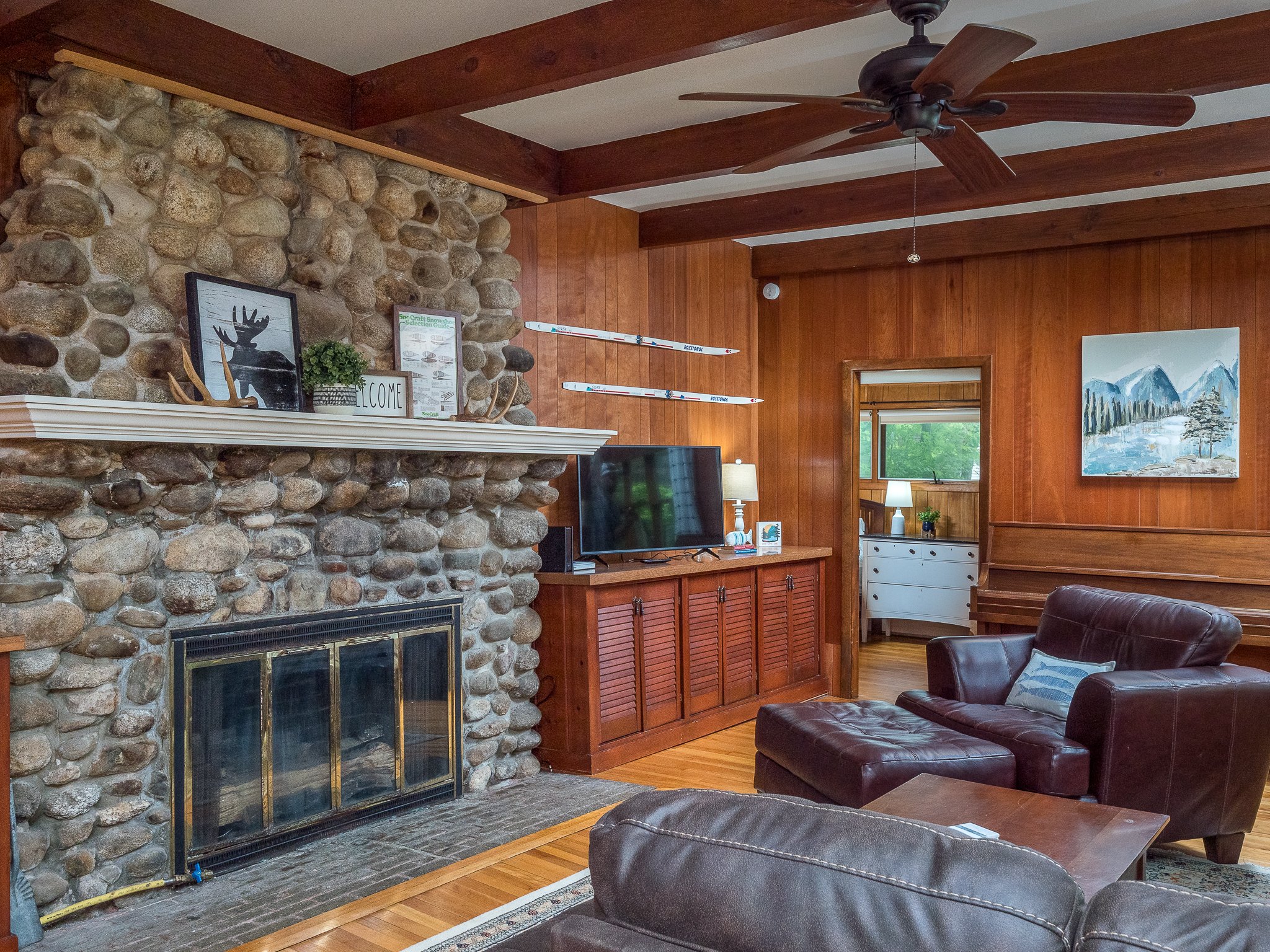 Cozy living room with stone fireplace, leather armchairs, wooden walls, ceiling fan, and a TV on a cabinet. Artwork and decor items are on the fireplace mantle.