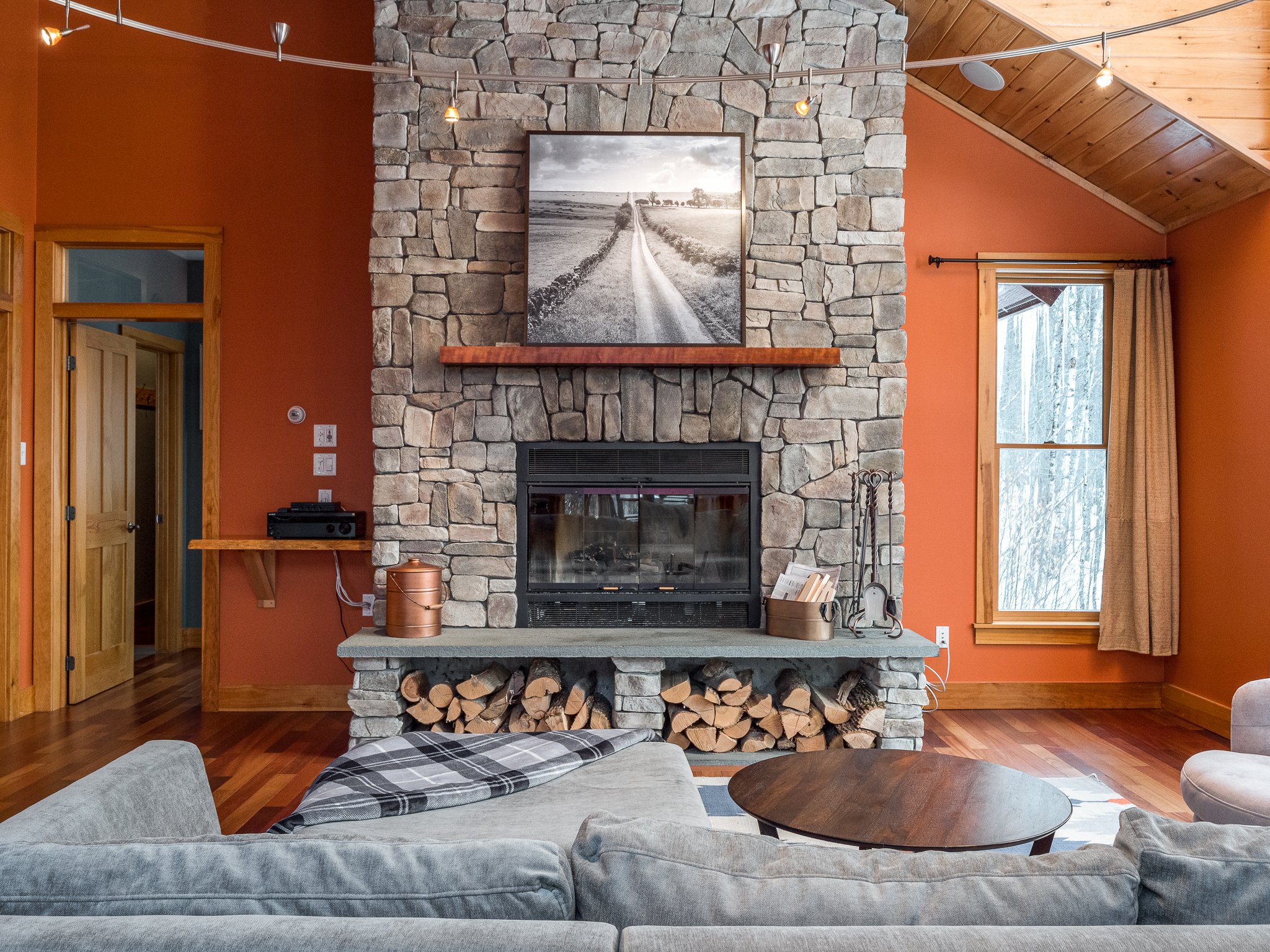 Cozy living room with stone fireplace, wood logs, gray sofa, and orange walls.