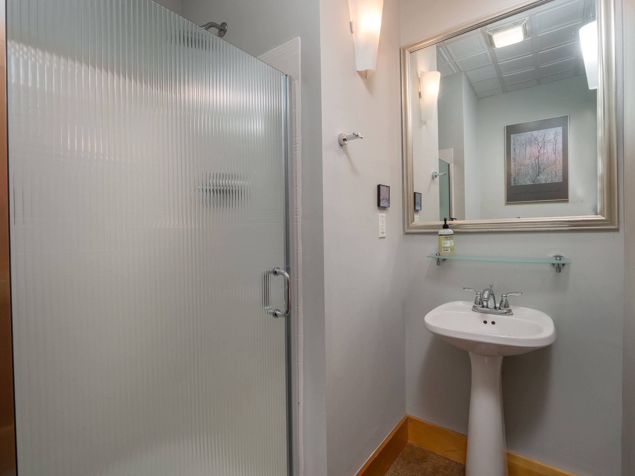 Modern bathroom with glass shower, pedestal sink, wall-mounted mirror, and wall light fixtures.