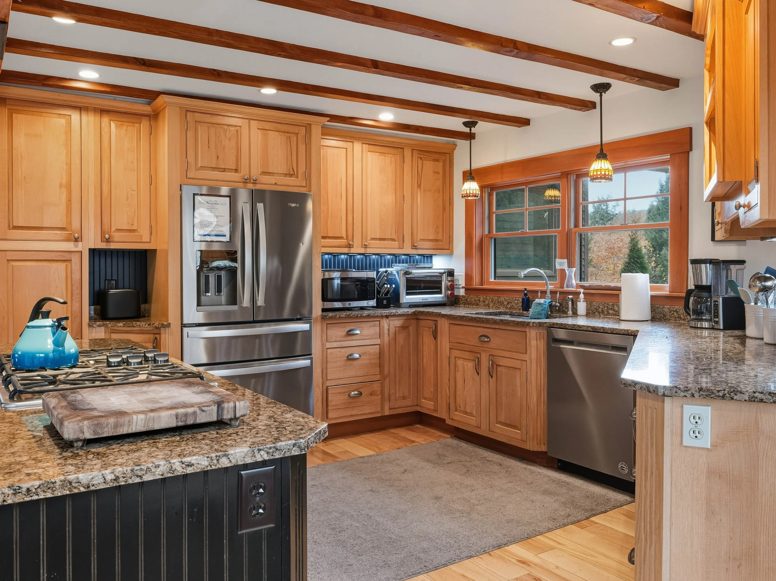 A kitchen with wooden cabinets, granite countertops, and stainless steel appliances, including a refrigerator and dishwasher. There are two hanging pendant lights over the window and a double-basin sink. The kitchen has wood beams on the ceiling and 