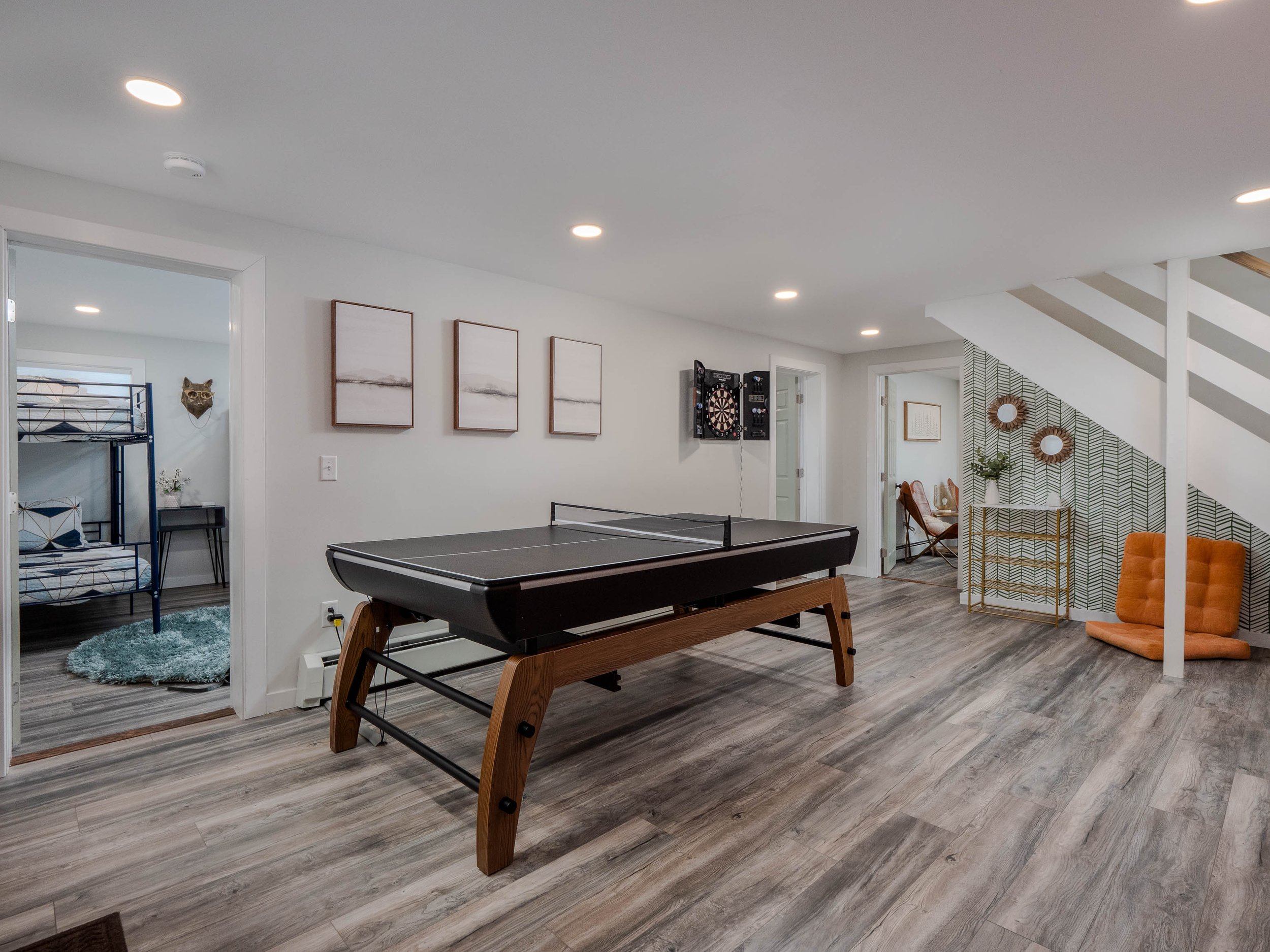 A spacious game room with a ping pong table, a dartboard, and seating areas, including an orange cushion and a velvet ottoman, with a patterned accent wall under a staircase.