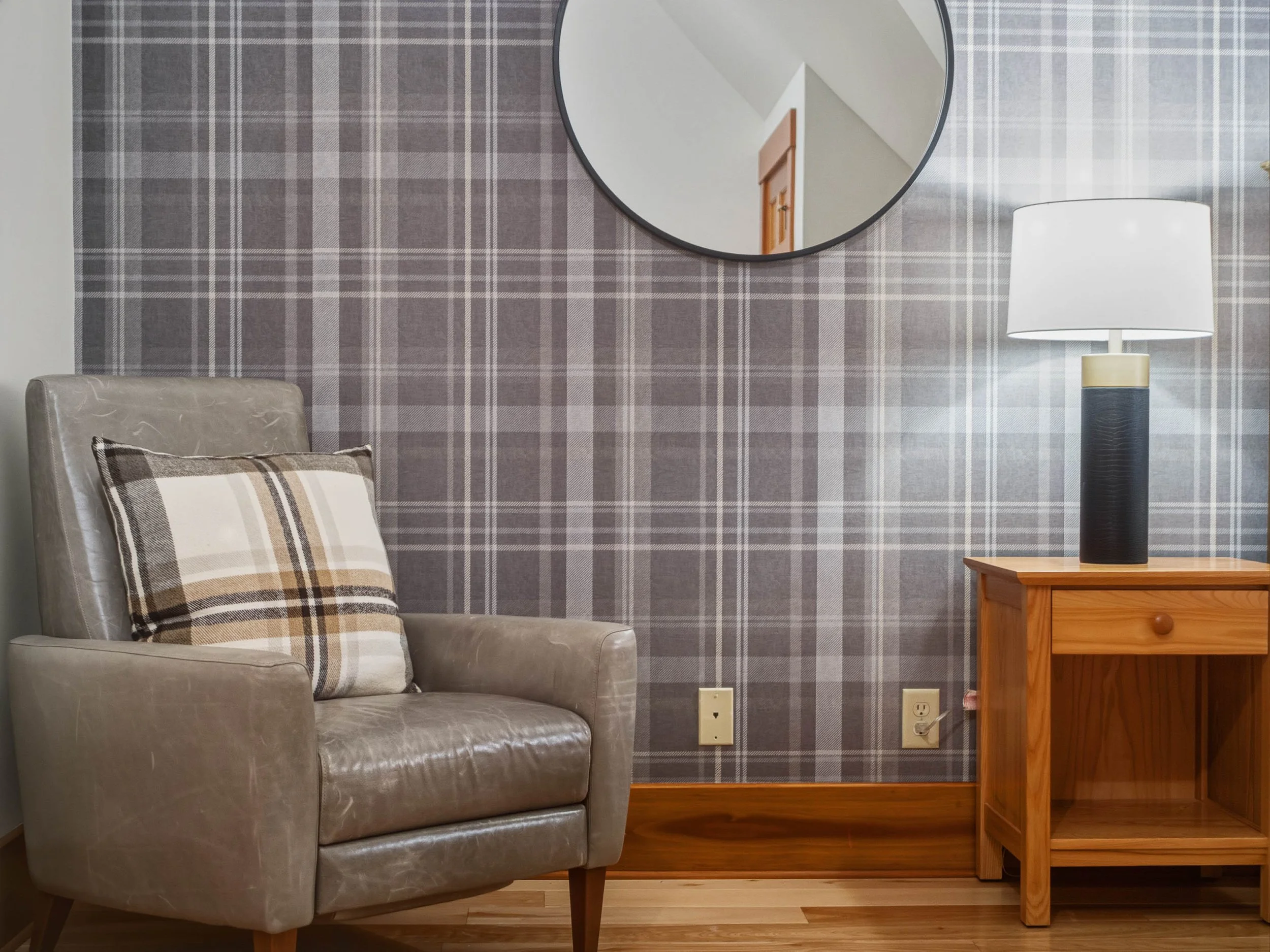 A living room corner with a beige leather armchair, a plaid cushion, a wooden side table with a tall black and white table lamp, a wallpapered wall with a round mirror, and multiple electrical outlets.