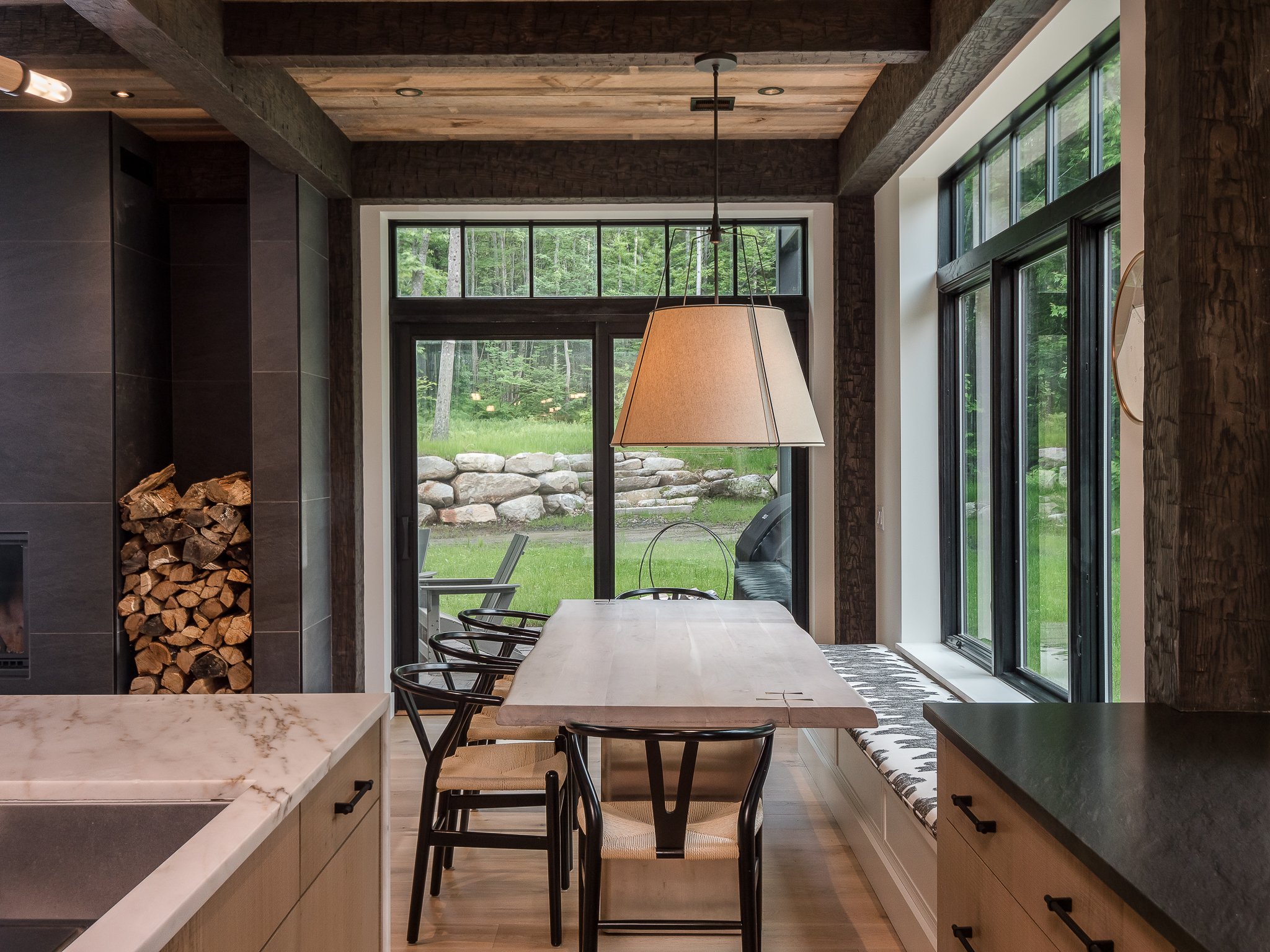 Modern dining area with a wooden table, black chairs, large pendant lamp, and windows overlooking a green yard; firewood stacked beside a grey wall.