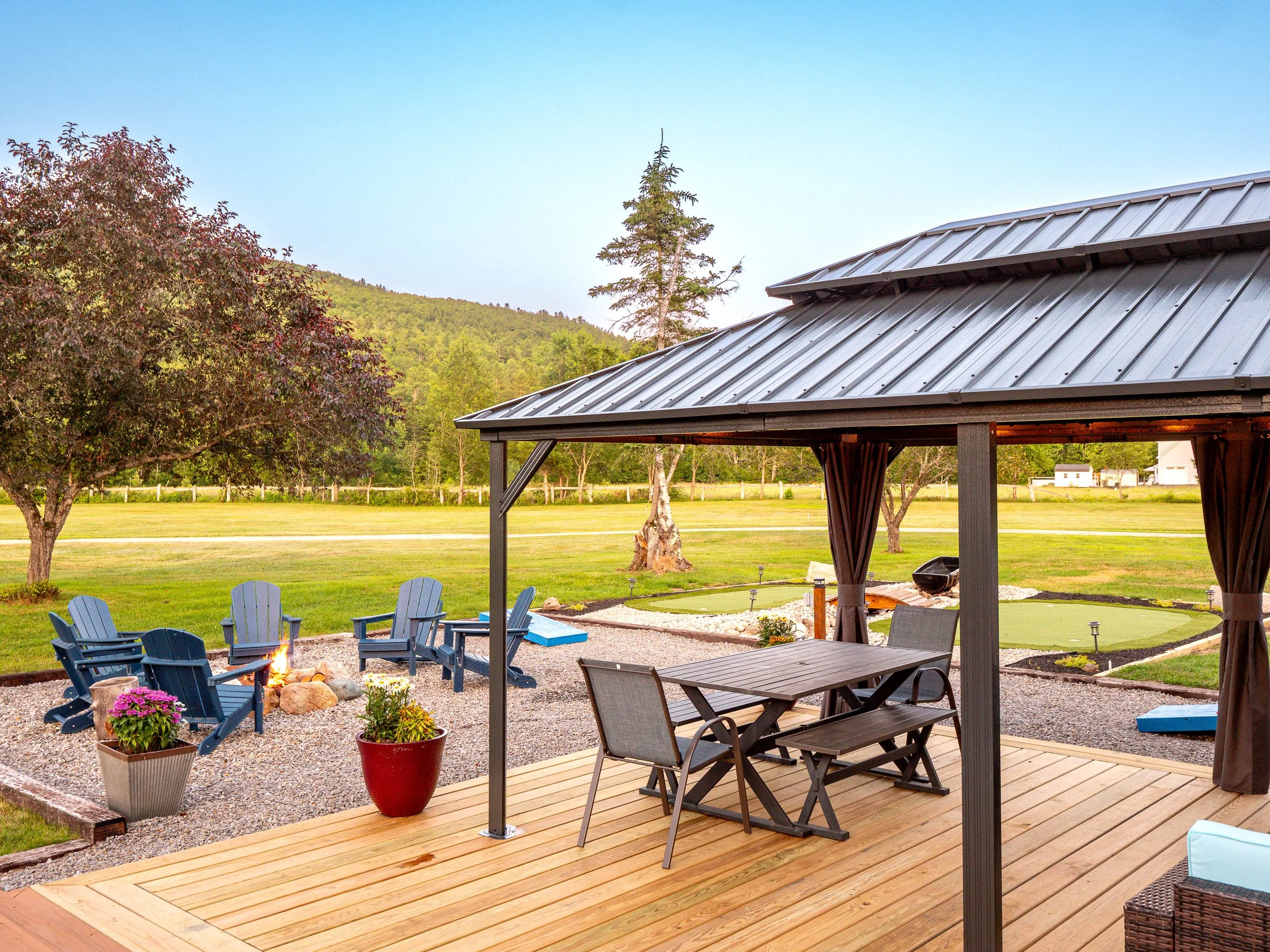 Backyard patio with a wooden deck, outdoor furniture, a fire pit with chairs, potted plants, and a well-manicured lawn with trees and a mountain in the background.