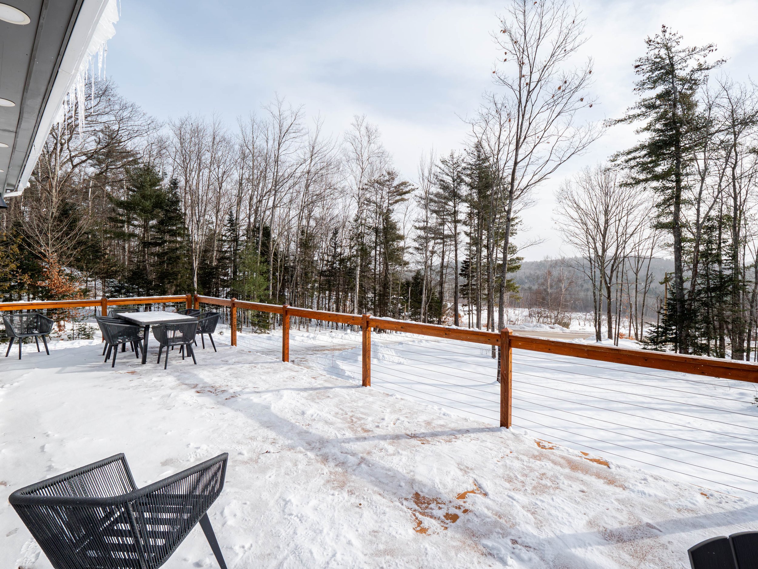 Snow-covered outdoor patio with black chairs and a table, overlooking a snow-covered wooded landscape with leafless trees and evergreens.