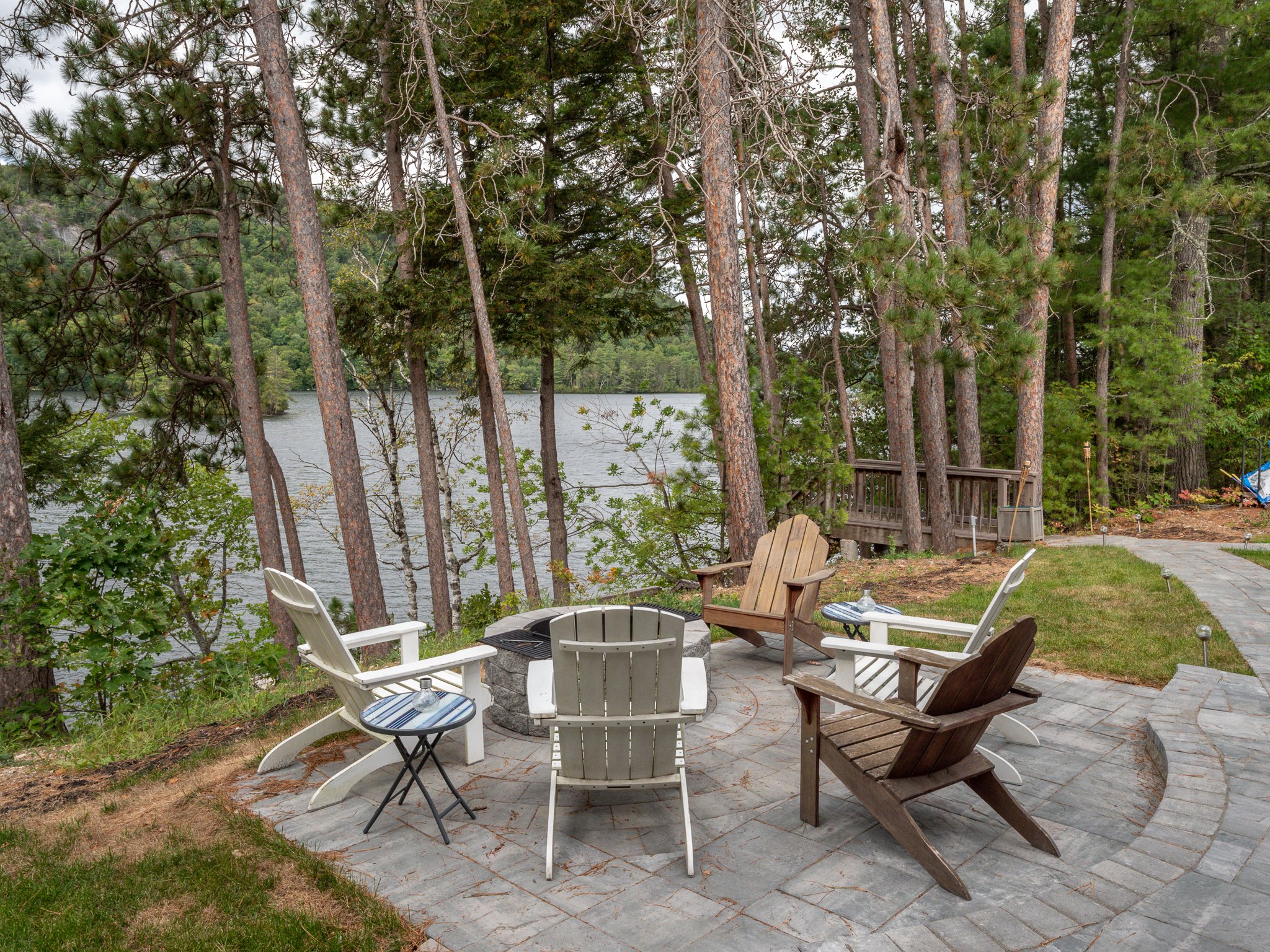 Outdoor seating area with Adirondack chairs around a fire pit, overlooking a lake surrounded by trees.
