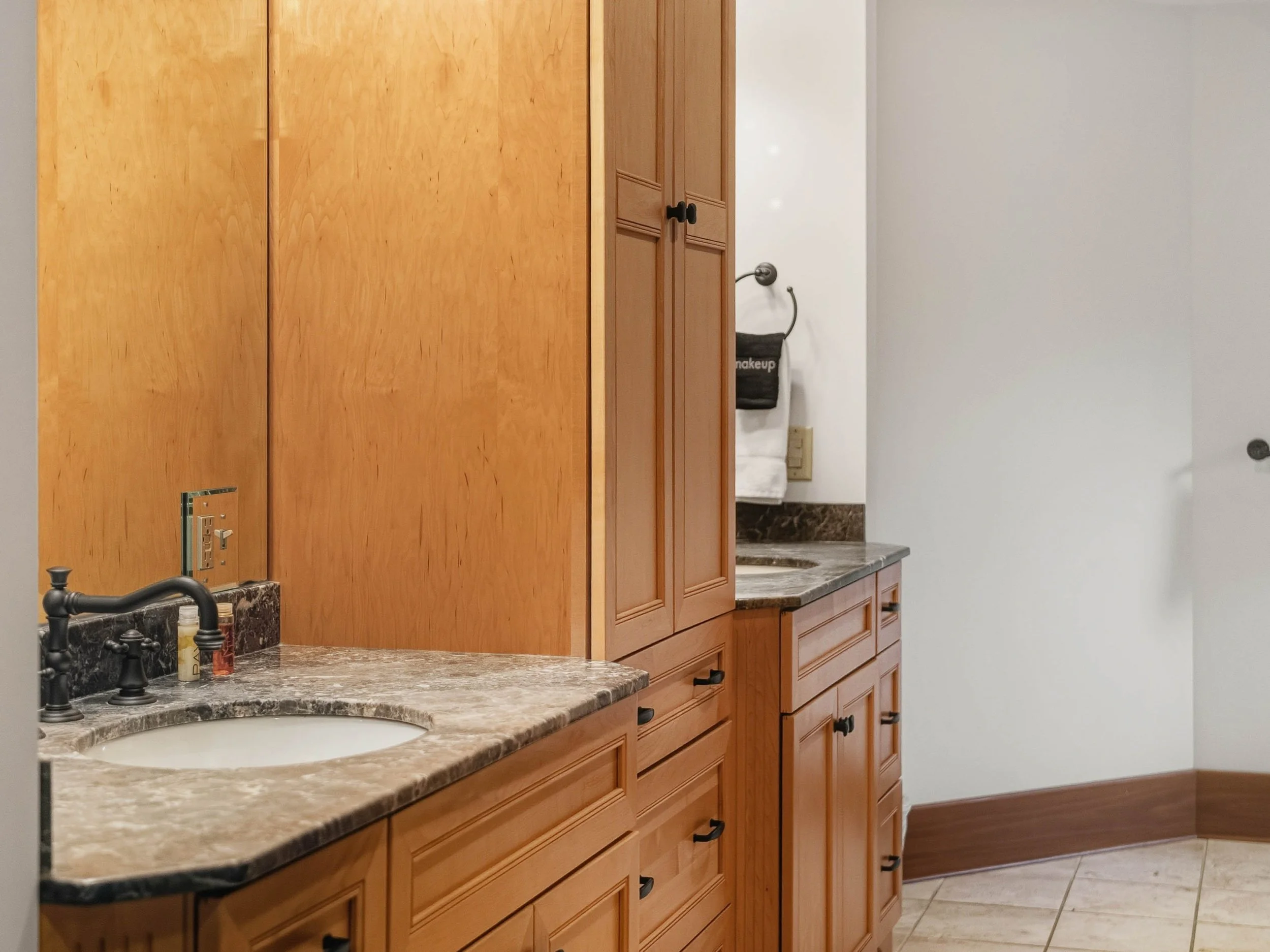 Bathroom with wooden cabinetry, marble countertops, and a white wall. A sink with a black faucet, a towel and a makeup bag hanging on a rack are visible.