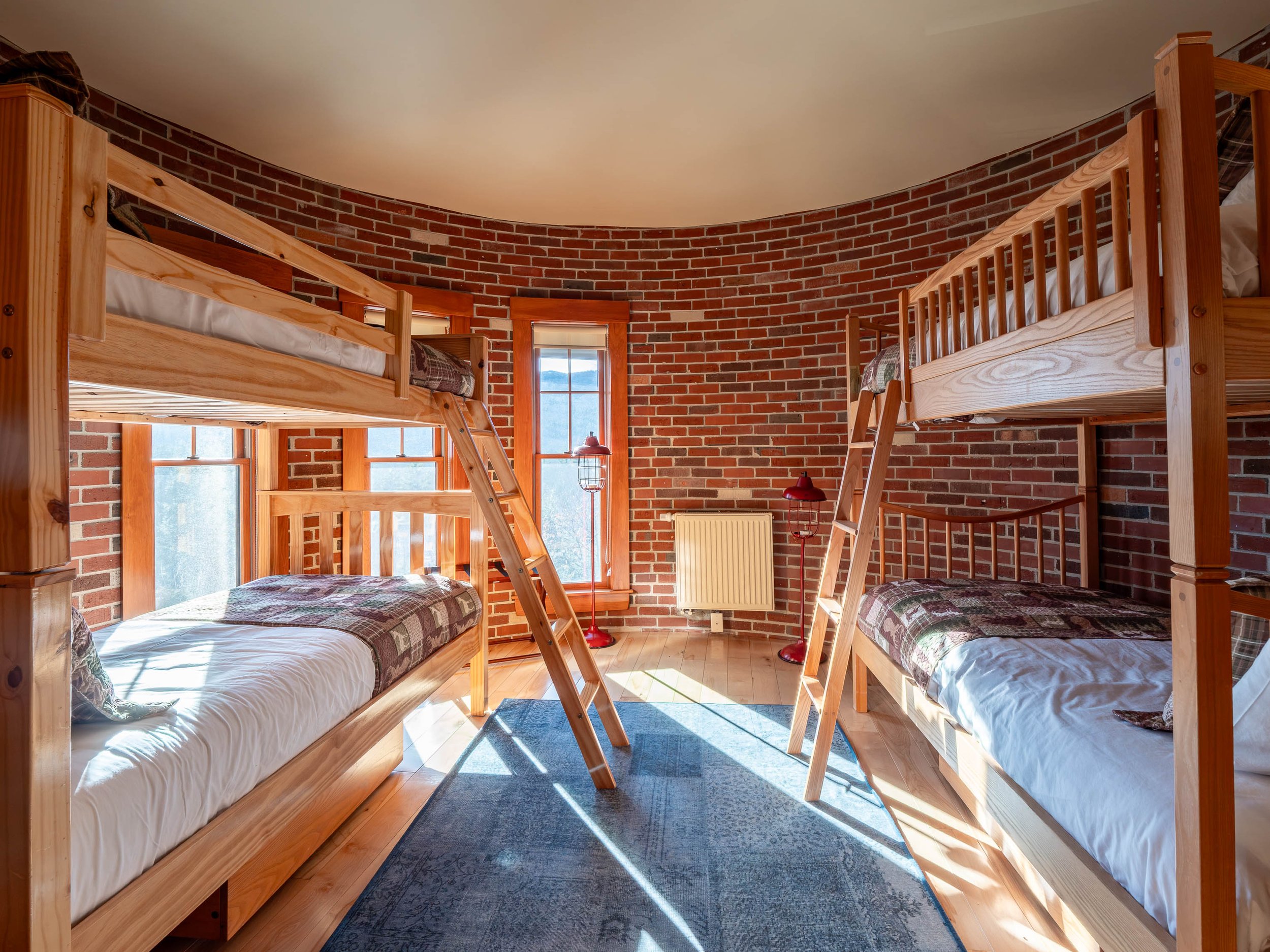 A bedroom with brick walls, wooden floors, and two bunk beds on either side, each with a ladder, white bedding, and plaid blankets. There are three windows with wooden frames, a blue area rug, two red floor lamps, and a radiator on the back wall.