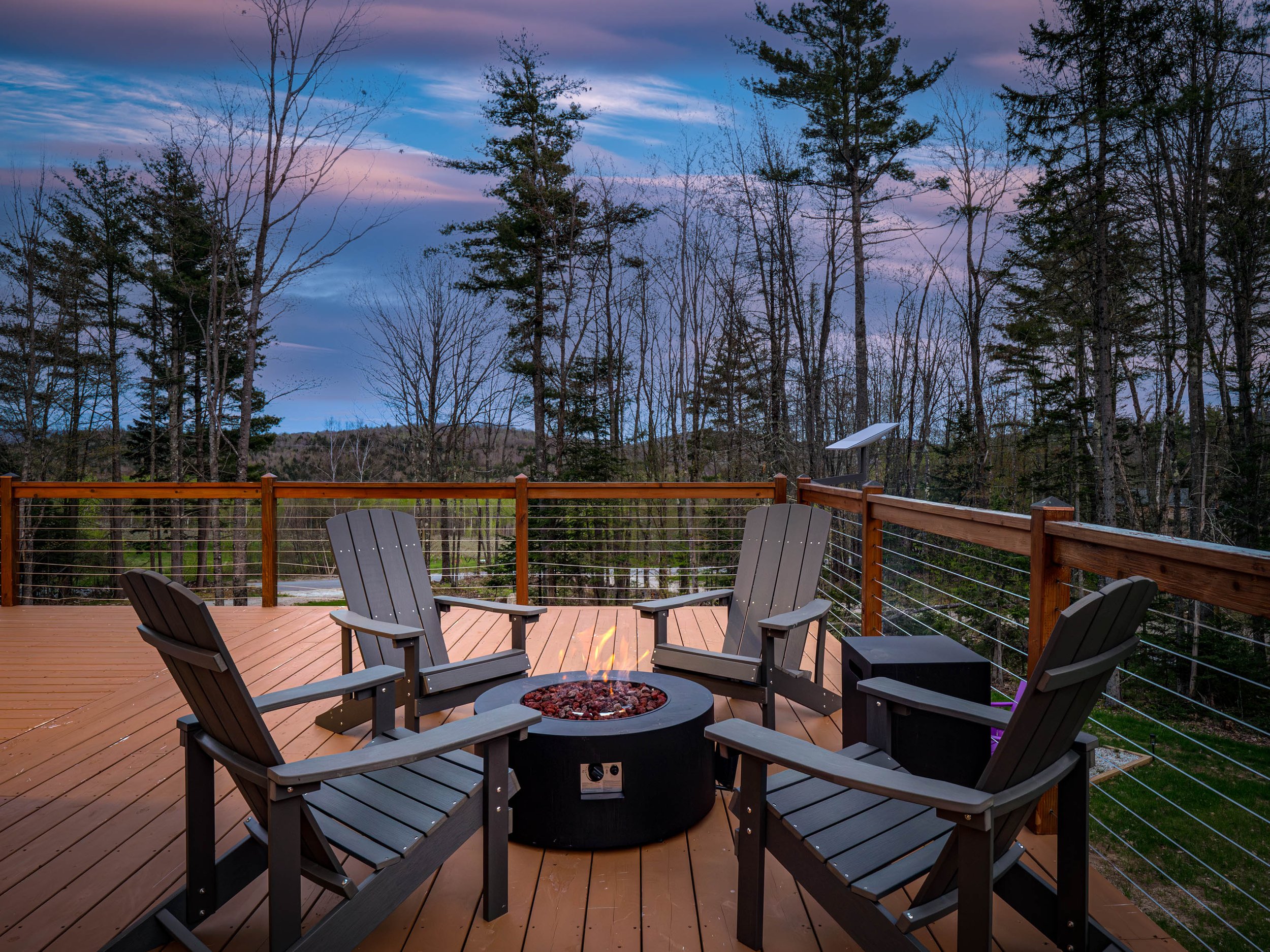 A wooden outdoor deck with four Adirondack chairs arranged around a fire pit, overlooking a forested area with tall trees at dusk.