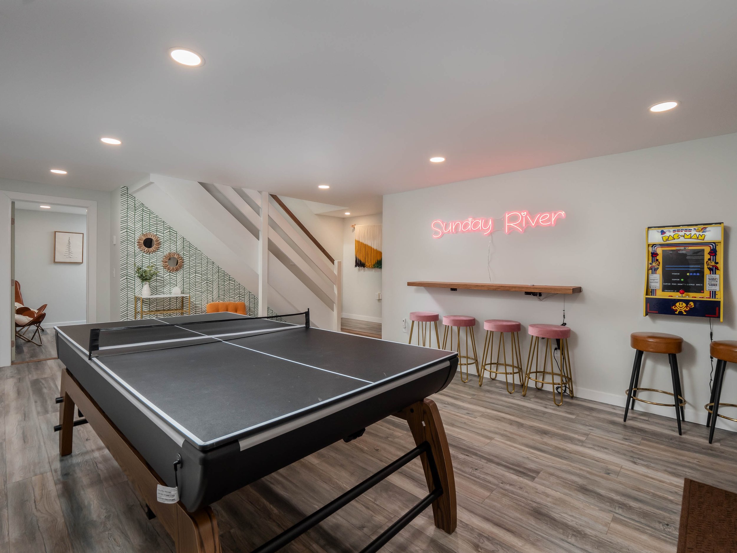Indoor game room with ping pong table, neon sign reading 'Sunday River', arcade game, staircase, and bar stools.