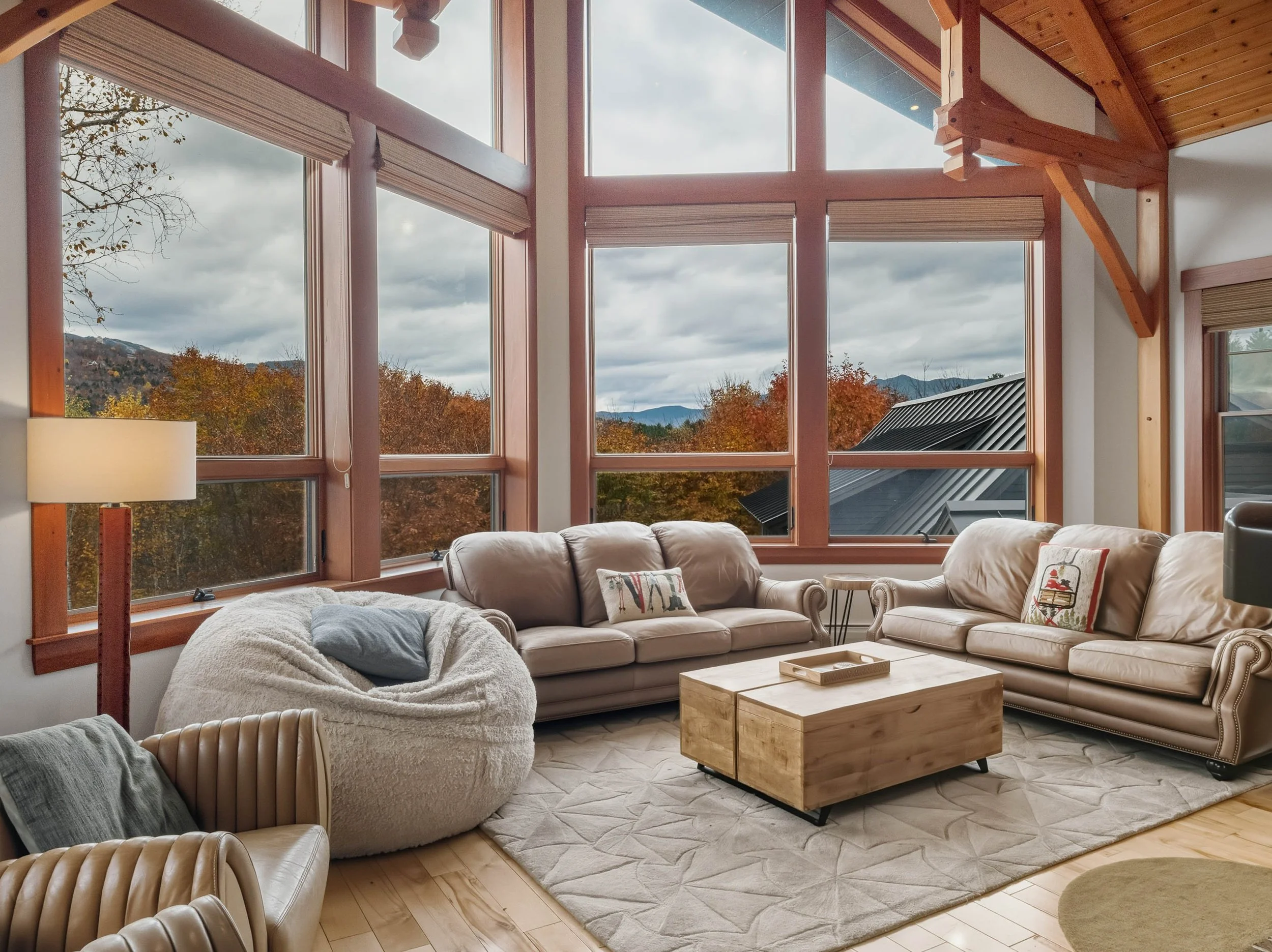 Living room with large windows showing trees and mountains, beige sofas, a cream armchair with a pillow, a wooden coffee table, a beige rug, and a lamp.