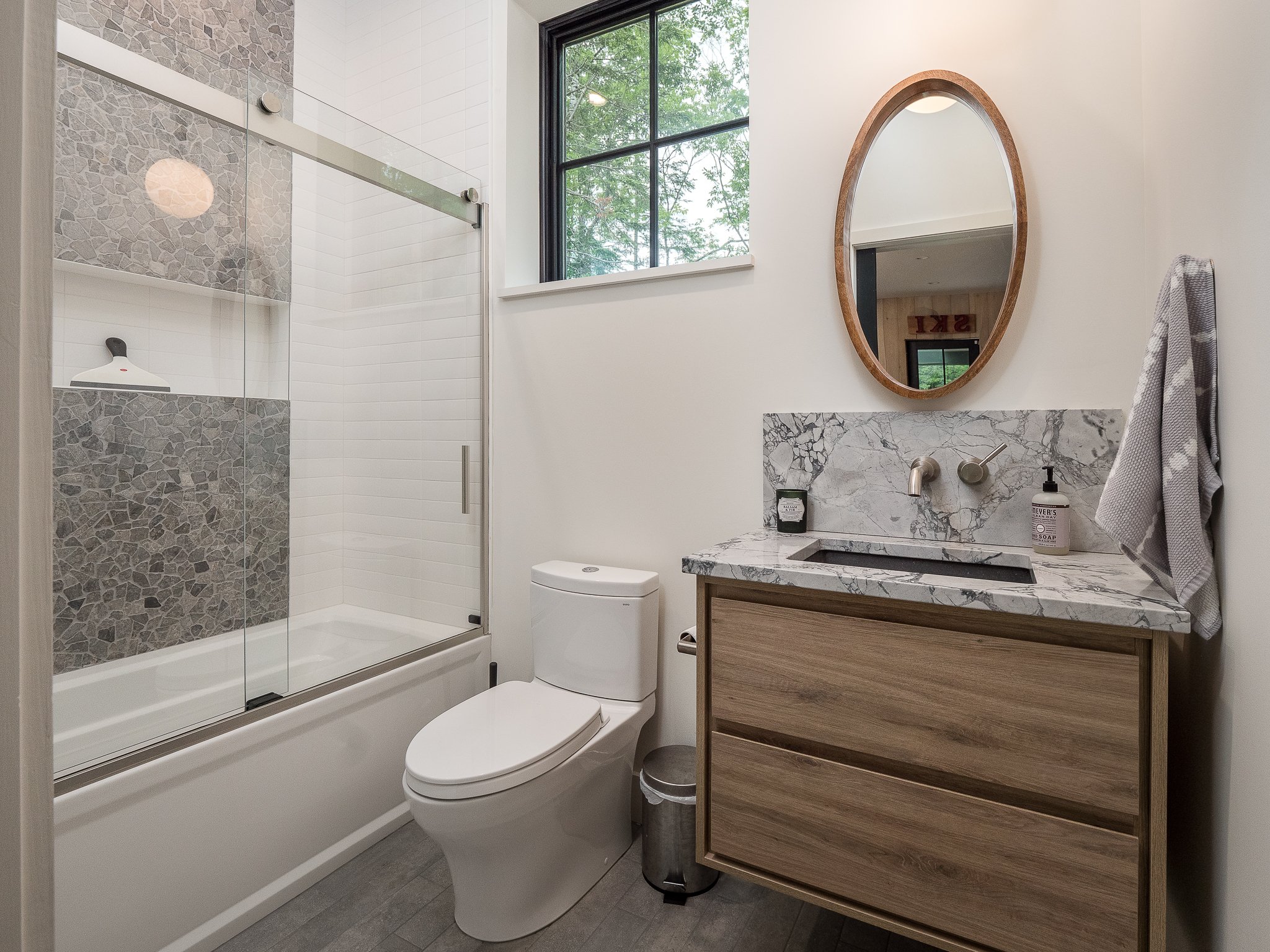 Modern bathroom with glass shower, white toilet, wooden vanity, marble countertop, oval mirror, and towel on wall hook.