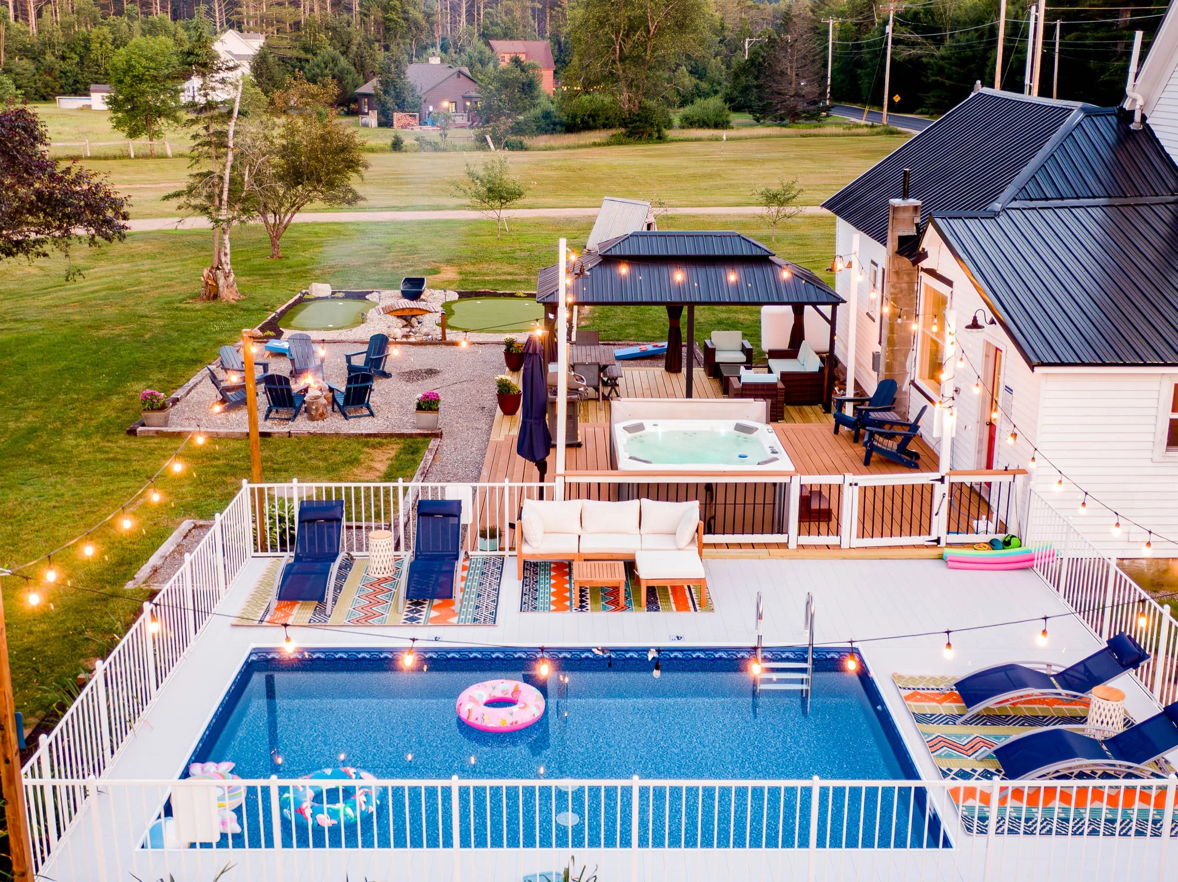 Backyard with a swimming pool, lounge chairs, outdoor seating, hot tub, and a fire pit area in a grassy yard, with string lights overhead.