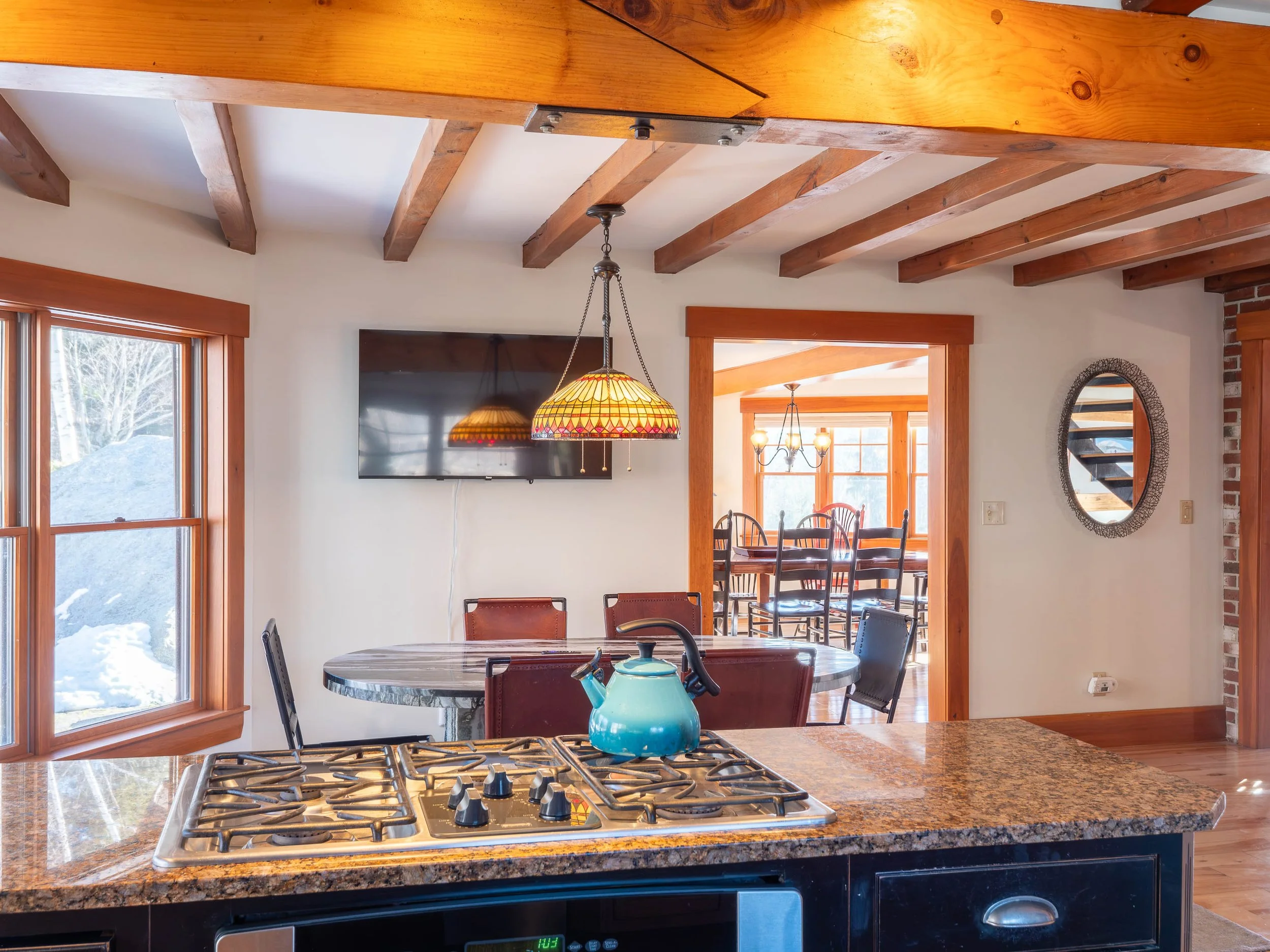 Kitchen with a granite island, a blue teapot, a wall-mounted TV, dining area with a round table and chairs, wooden beams on ceiling, and a window showing snow outside.