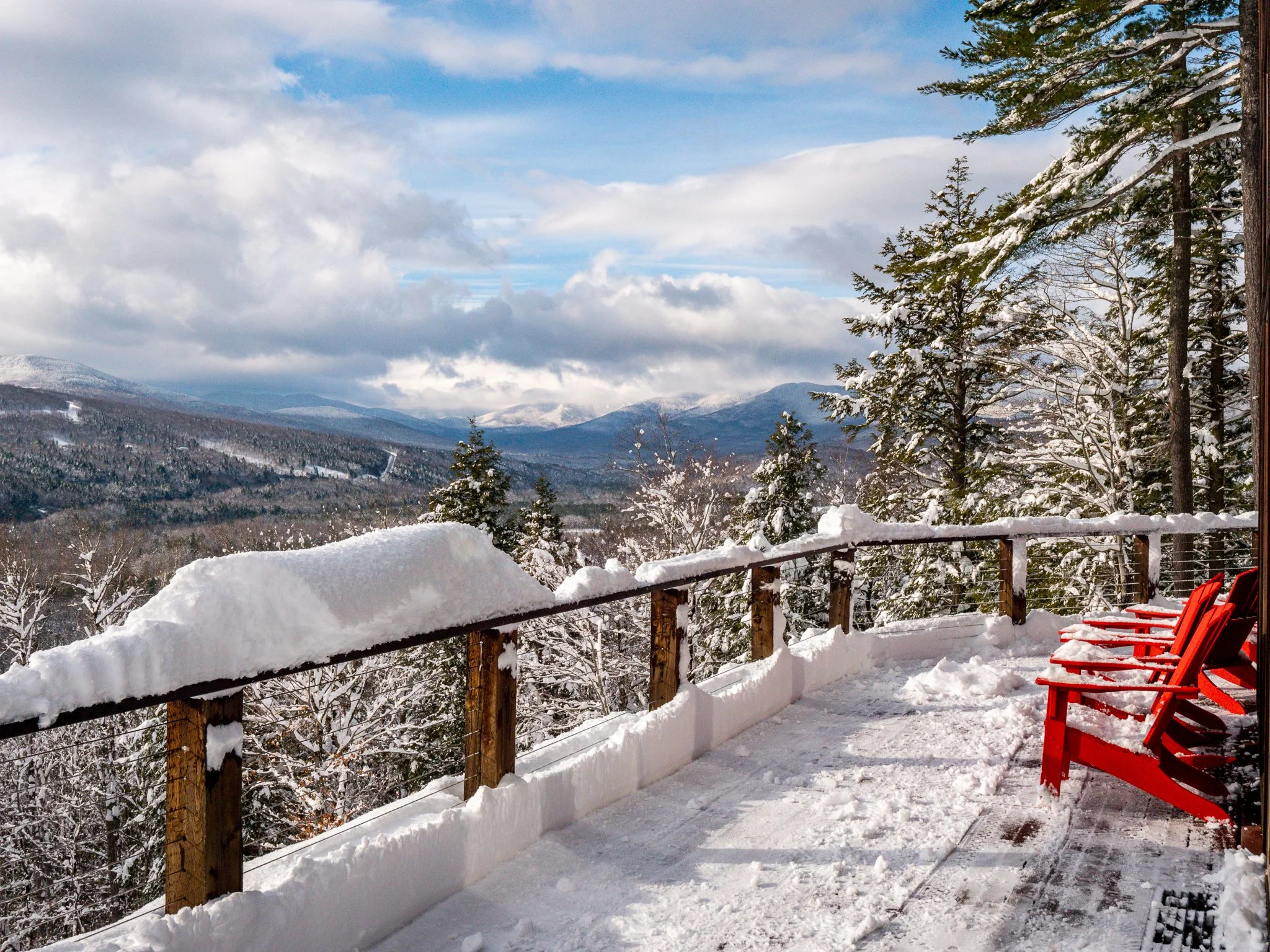 Snow-covered mountain landscape with a wooden deck and red Adirondack chairs in the foreground.