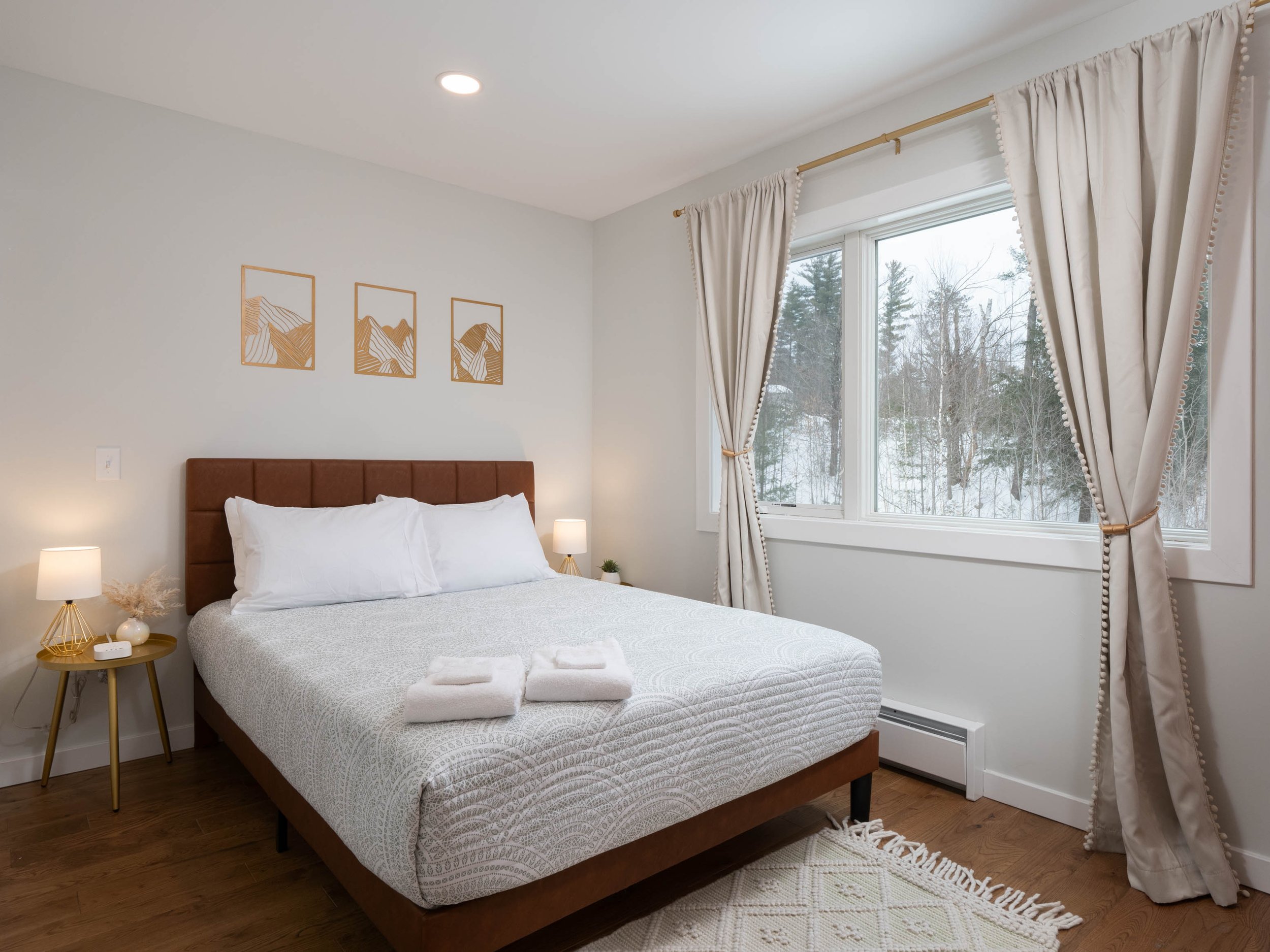 Clean bedroom with a large bed, white bedding, and two folded towels on top. Nightstands with lamps on each side and a window with beige curtains showing a snowy landscape outside. Wall art of mountain sketches above the bed.