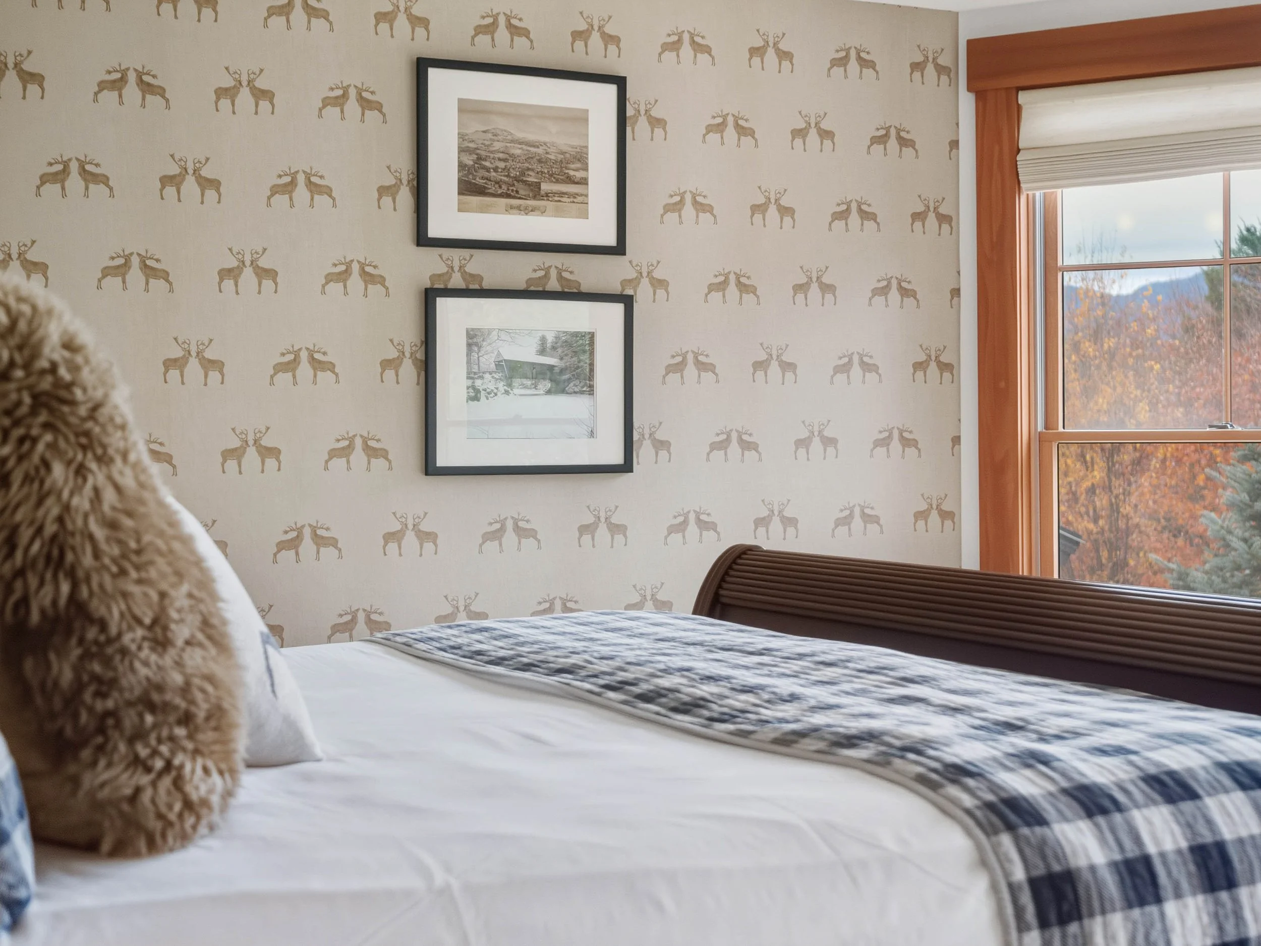 A bedroom with a bed, beige deer-patterned wallpaper, framed pictures on the wall, and a large window showing fall foliage outside.
