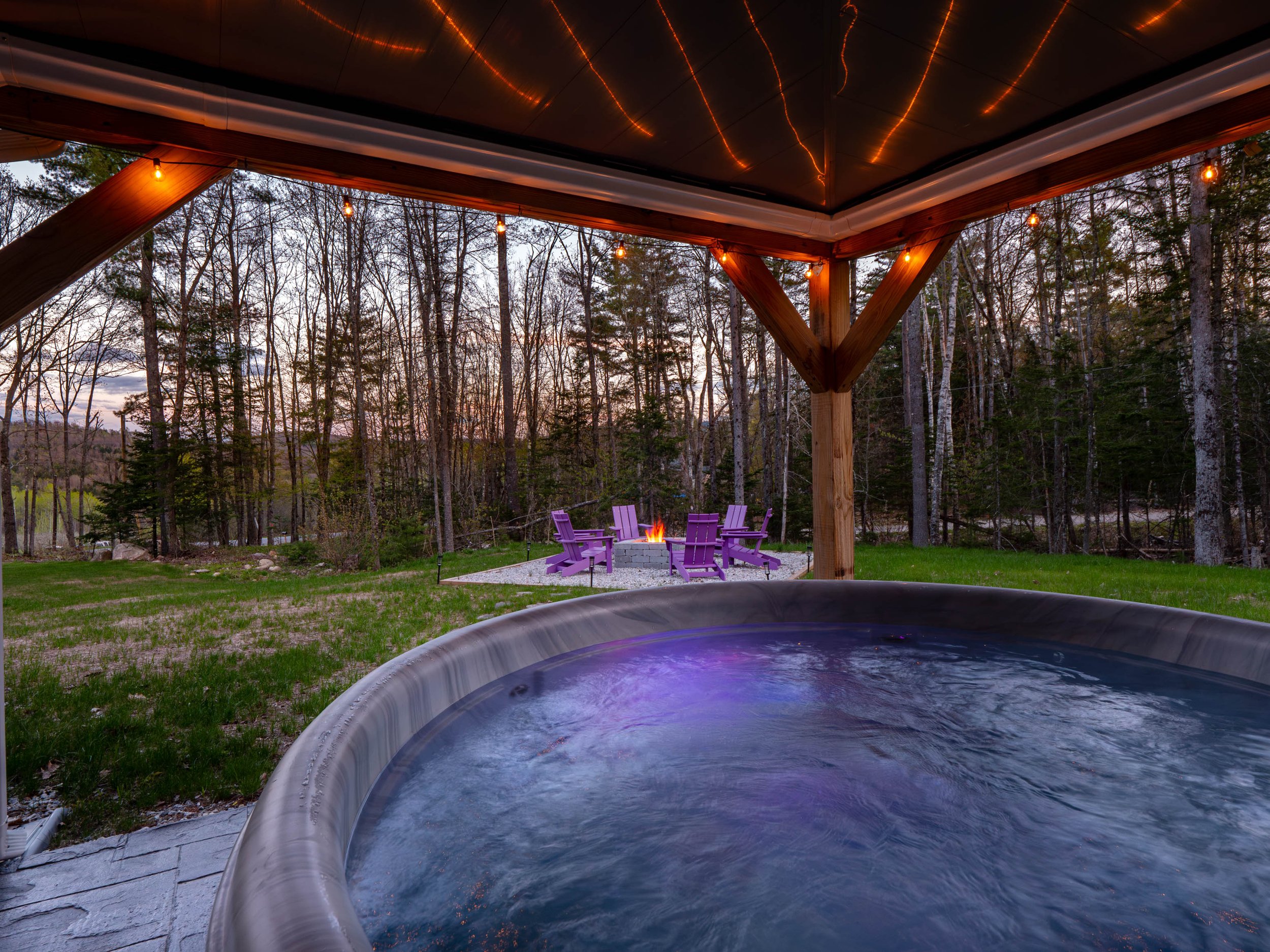 View of an outdoor hot tub under a wooden deck with string lights, overlooking a backyard with chairs around a fire pit, surrounded by trees at dusk.