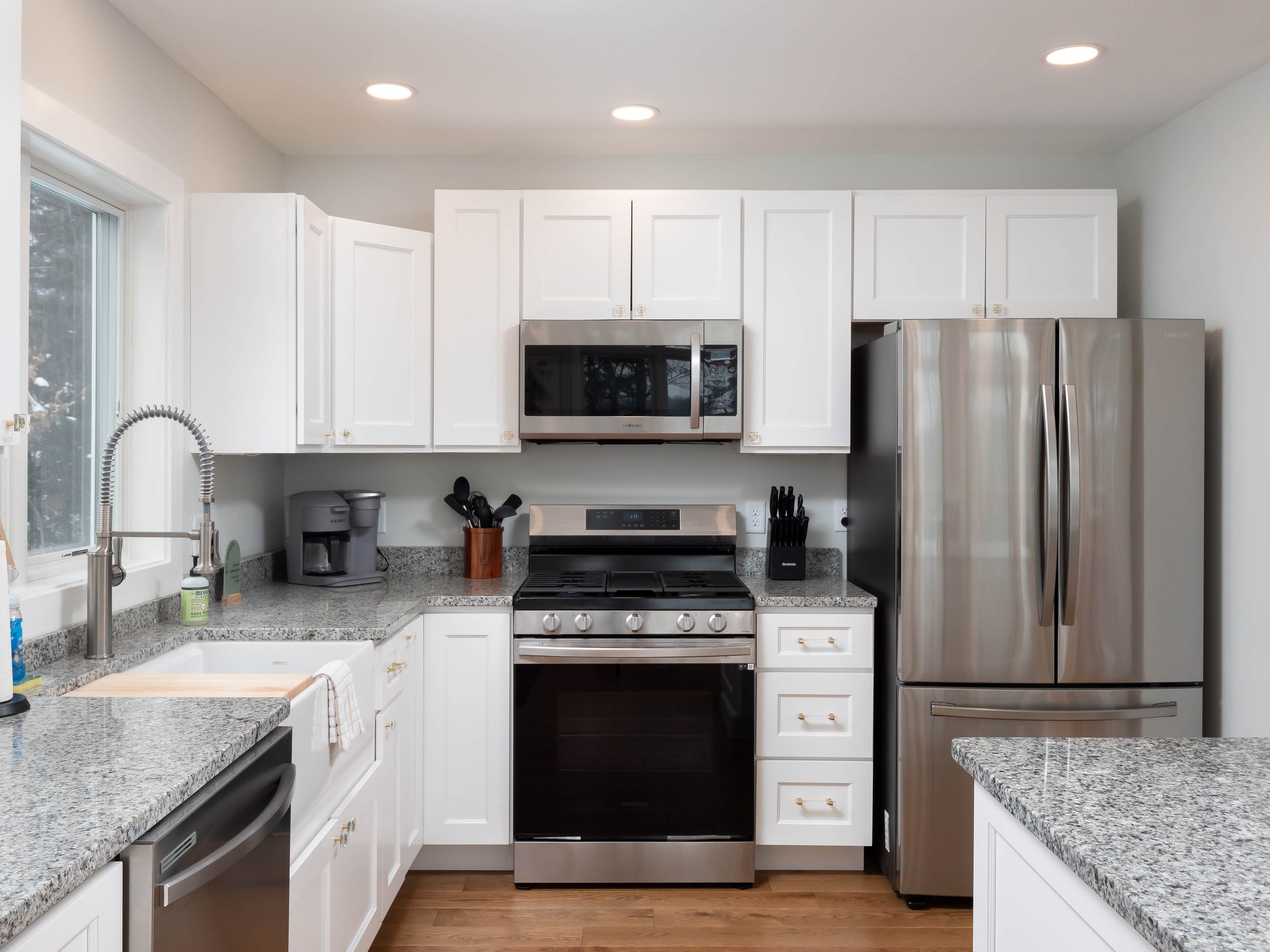Modern kitchen with white cabinets, stainless steel refrigerator and oven, microwave, granite countertops, and a window above the sink.