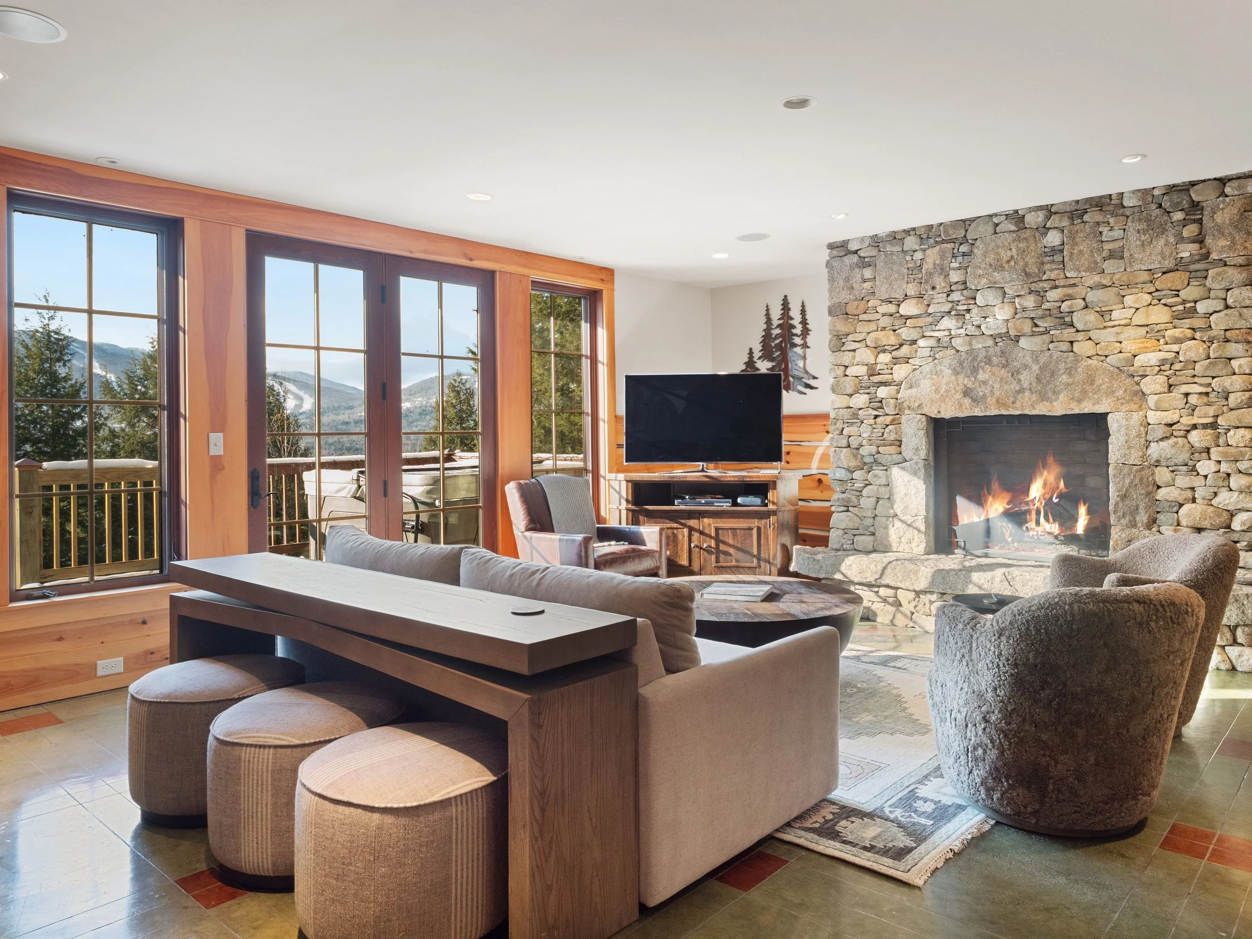 Living room with large windows, a stone fireplace with a fire, a flat screen TV, a beige sofa, a brown armchair, a wooden coffee table, three beige ottomans, and mountain scenery visible through the windows.