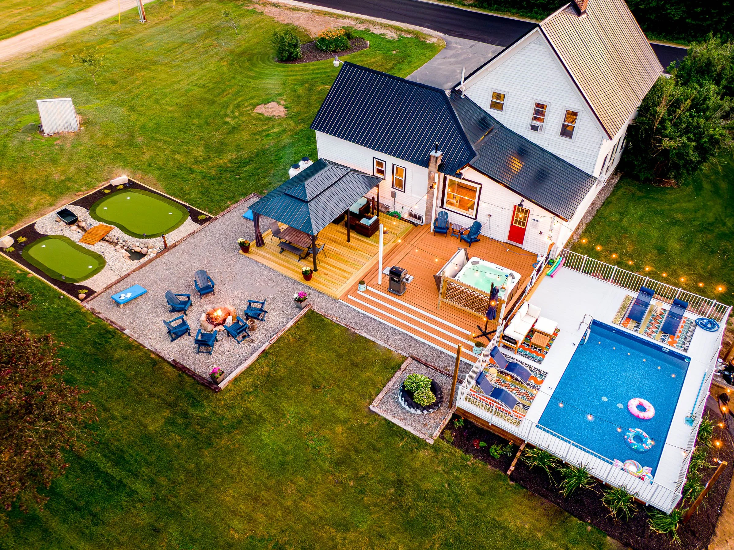 Aerial view of a backyard with a multi-level deck, hot tub, swimming pool, fire pit with chairs, two small pond-like water features, and a house with a garden area.