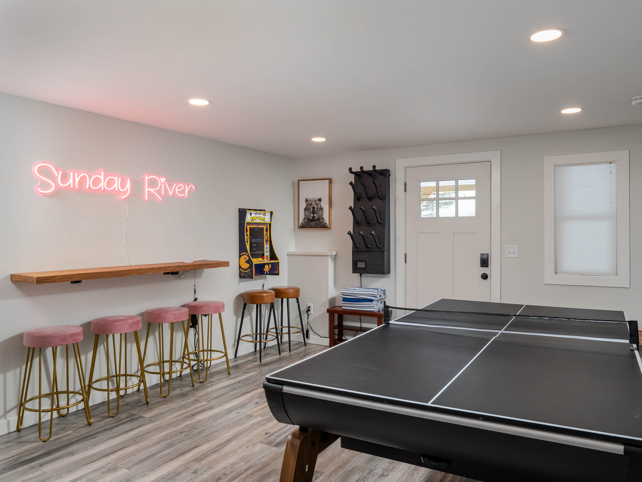 Game room with Ping-Pong table, bar stools, neon sign reading "Sunday River," arcade game, and black coat rack on white walls.