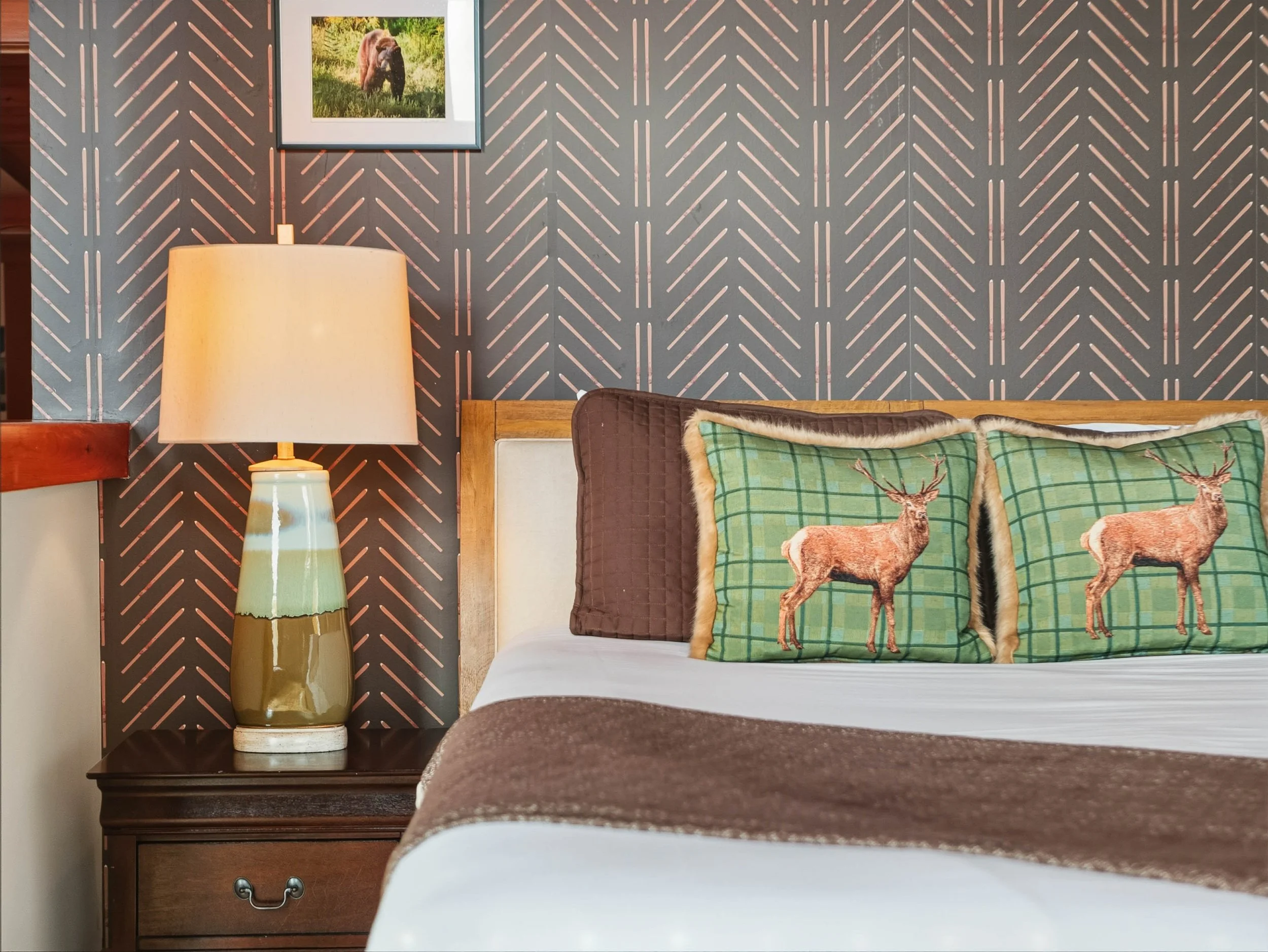 A bedroom with a bedside table, a lamp, and two pillows with deer designs. The wall has a patterned wallpaper with vertical lines and a framed photo of a bear.