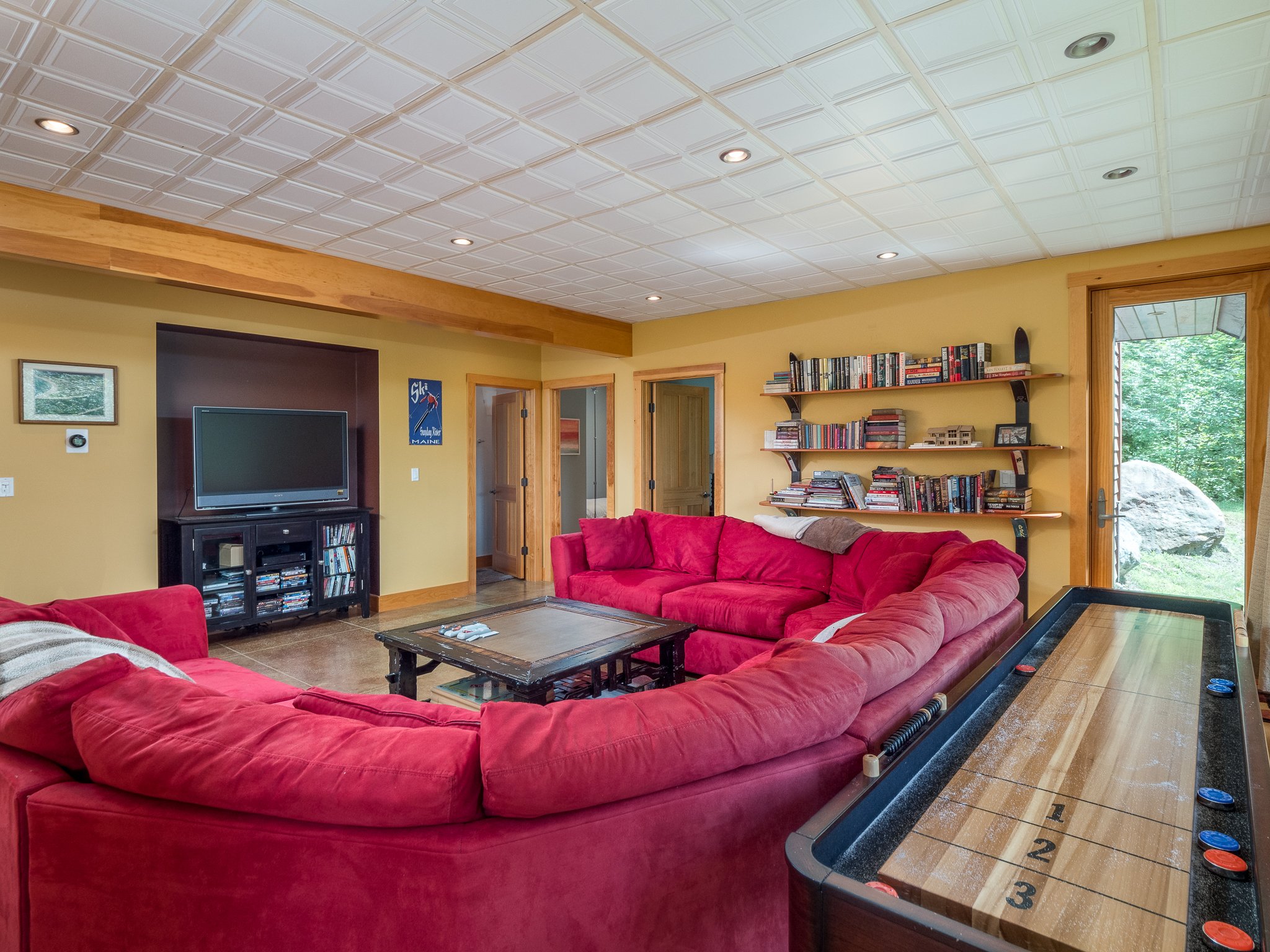 A cozy living room with red sectional sofa, TV, bookshelves, and shuffleboard table.