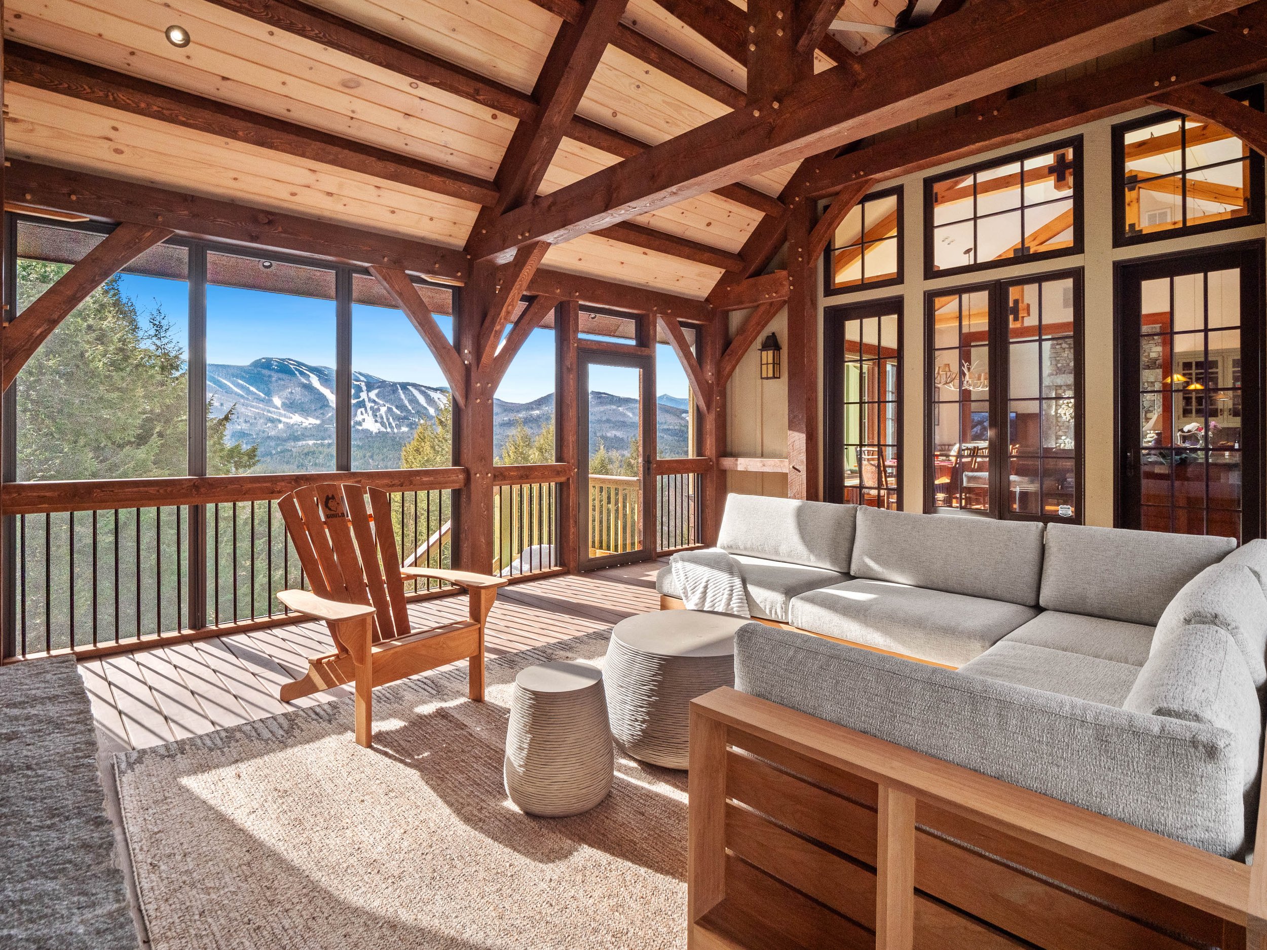 Living room with large windows displaying snow-capped mountains, featuring a beige sectional sofa, stone tables, a wooden chair, and a screened porch with a view.