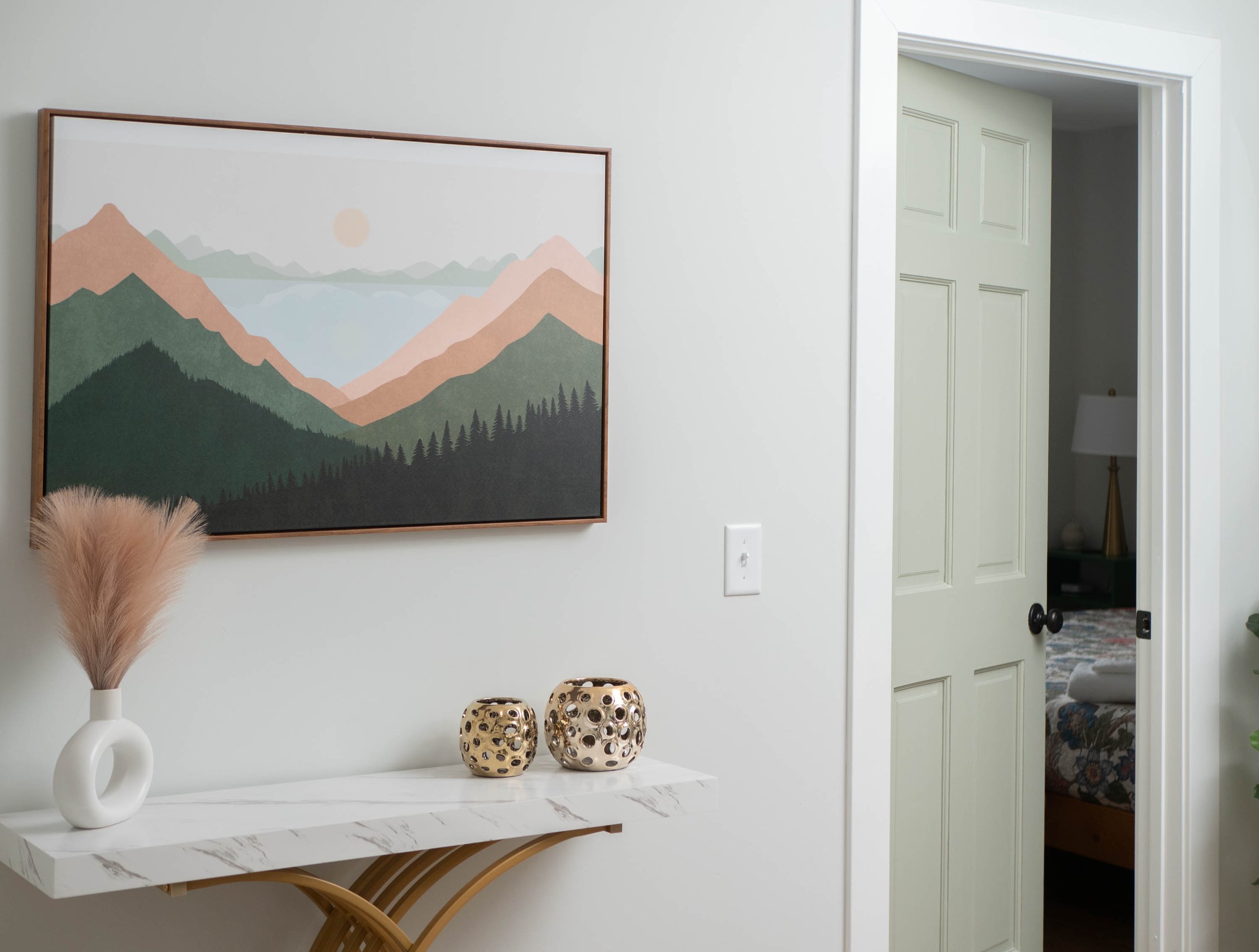 Decorative interior space with mountainous landscape artwork, a white console table with a vase and two gold candles, and a partially open door revealing a bedroom with a lamp and patterned bedspread.