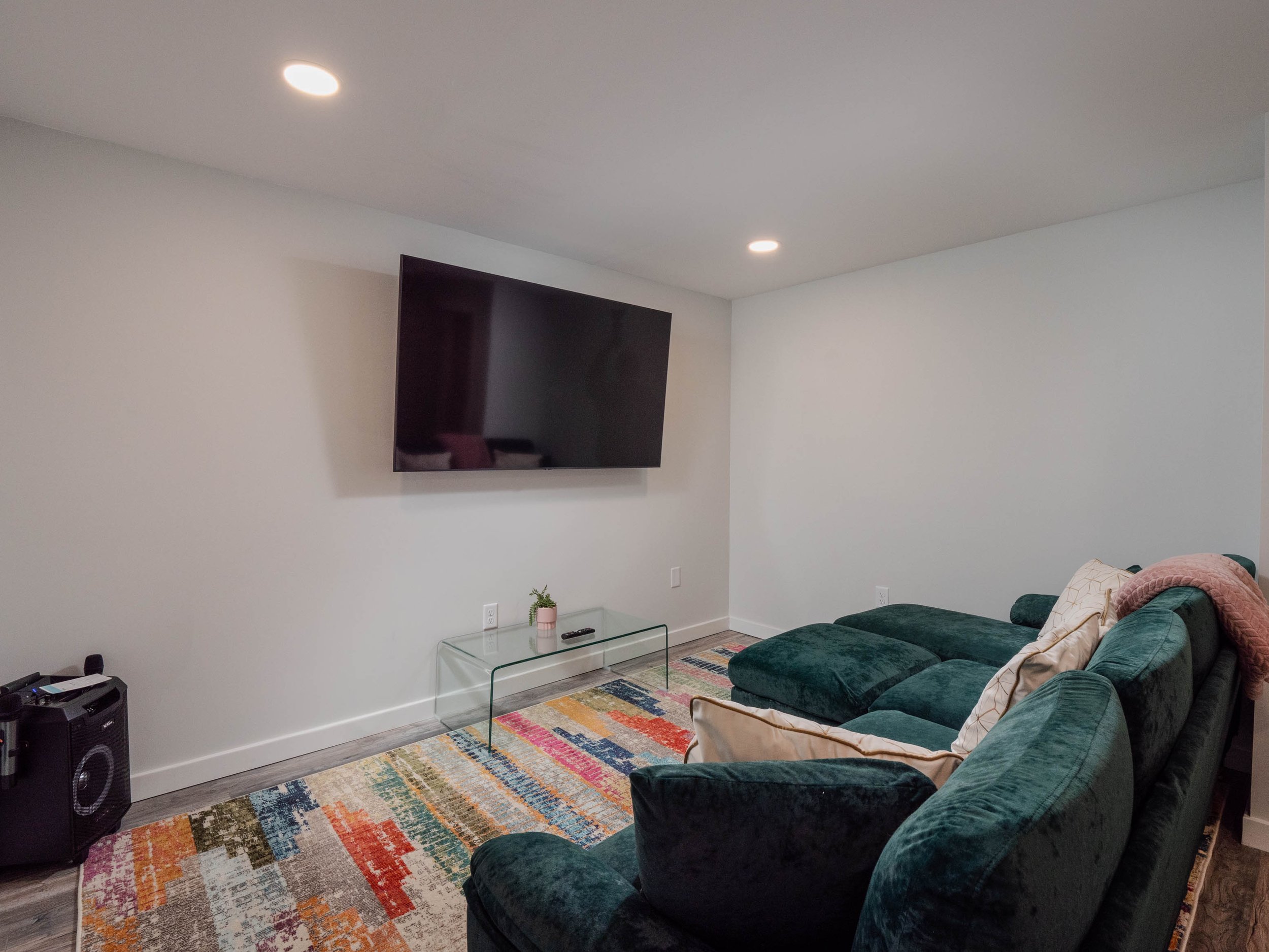 Living room with a large green sectional sofa, colorful area rug, mounted flat-screen TV on white wall, glass coffee table with a small potted plant and remote, and ceiling lights.