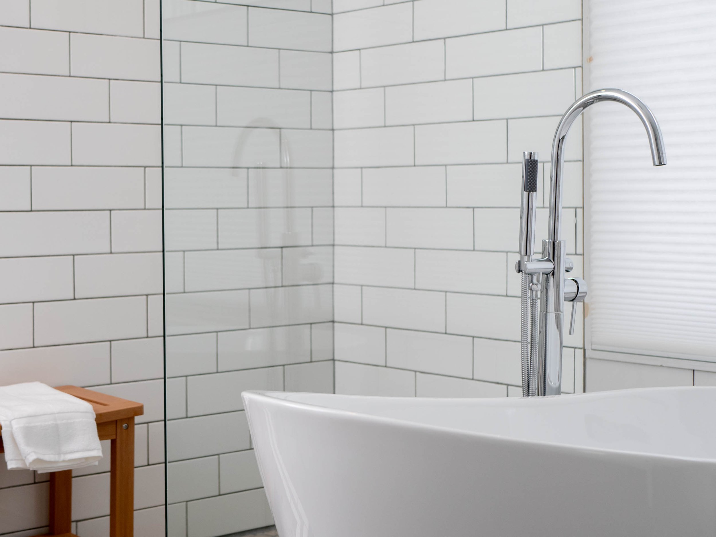 Modern bathroom with white subway tile walls, a white bathtub, a chrome shower fixture, and a small wooden side table with folded towels.