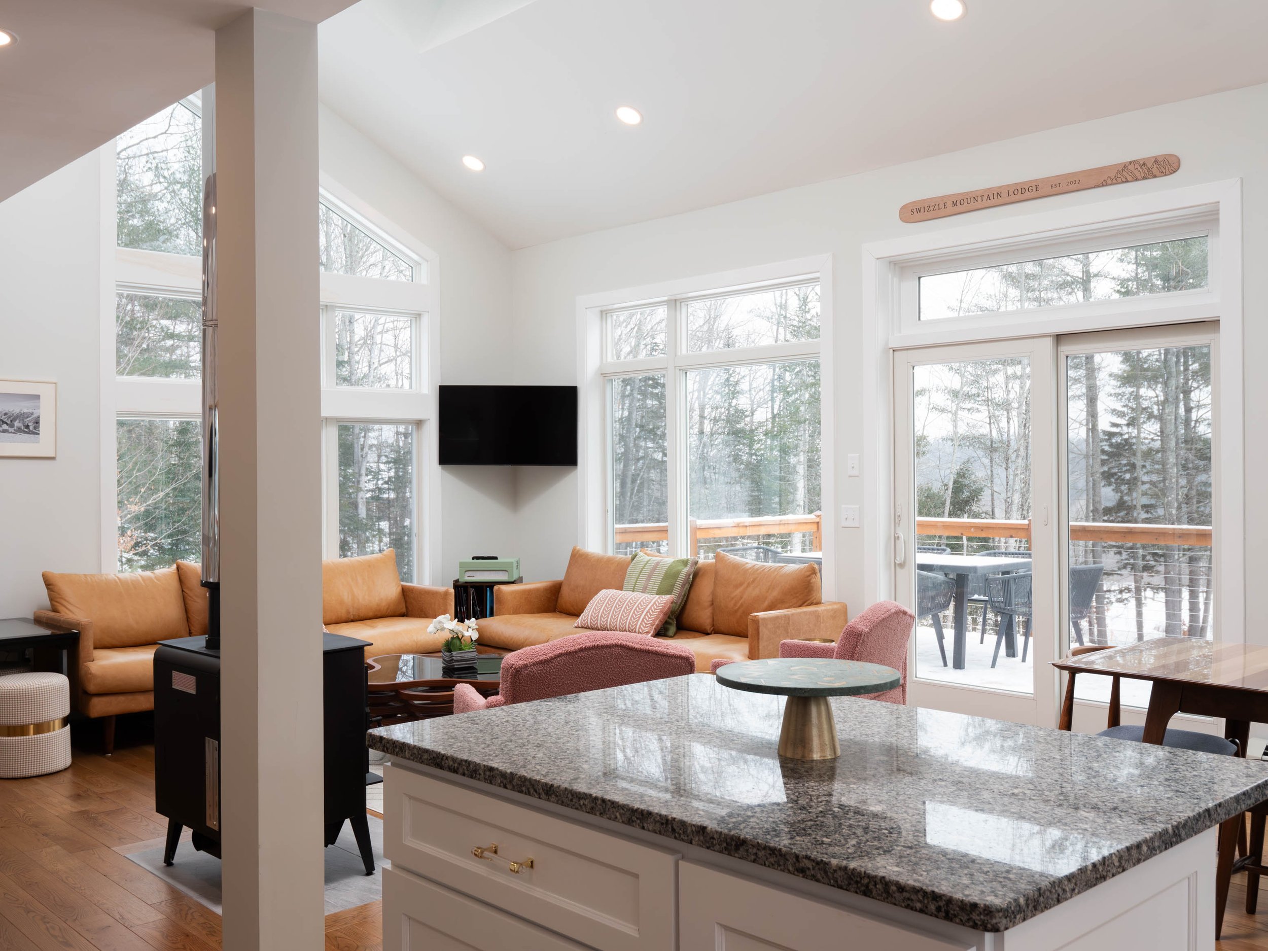 Living room with orange sofa, pink armchair, flat-screen TV, large windows showing a snowy outdoor deck, and a granite kitchen island with a decorative piece.