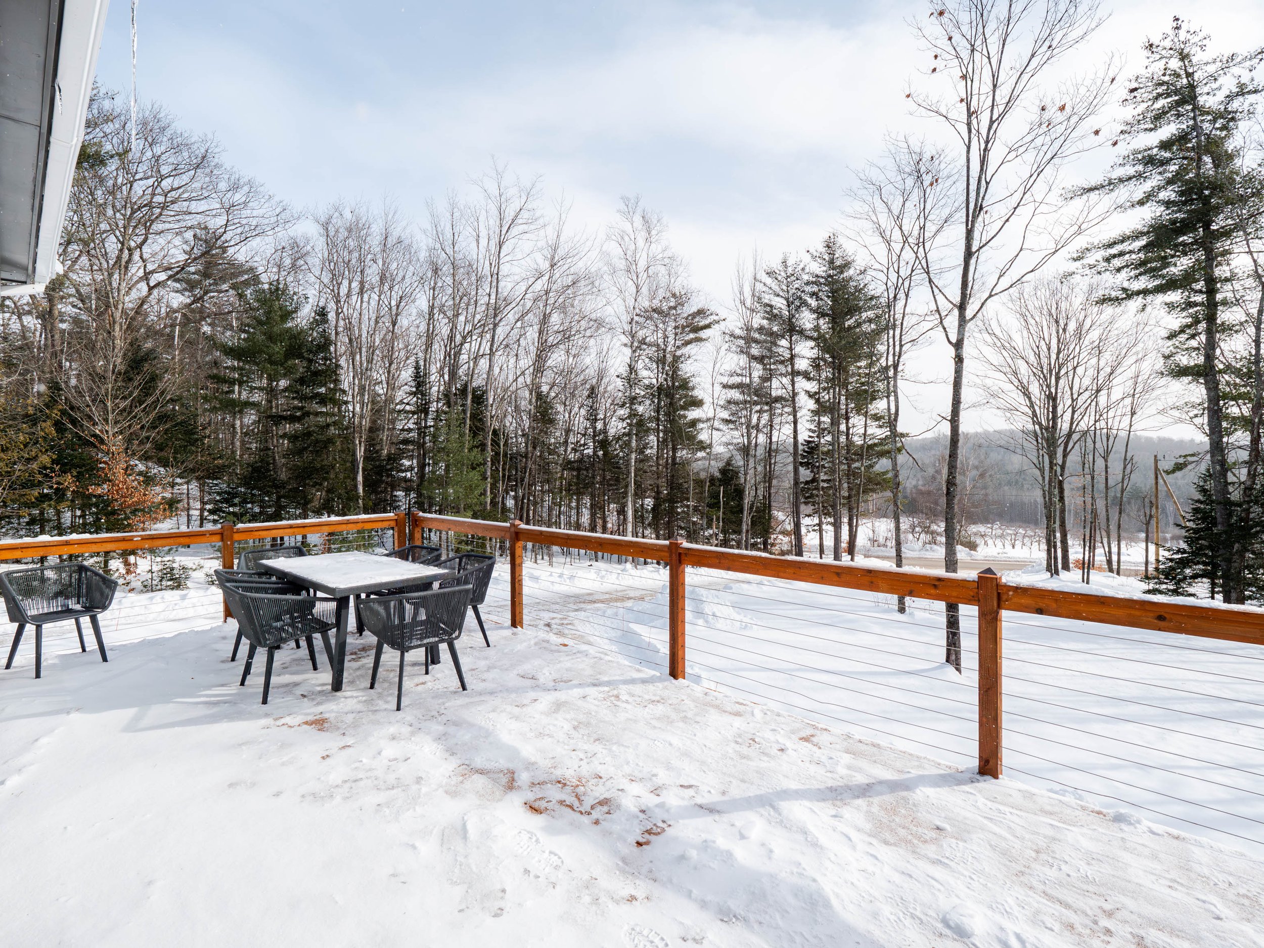 Snow-covered outdoor patio with black chairs and a table, surrounded by a wooden railing, overlooking a winter forest with bare trees and snow on the ground.
