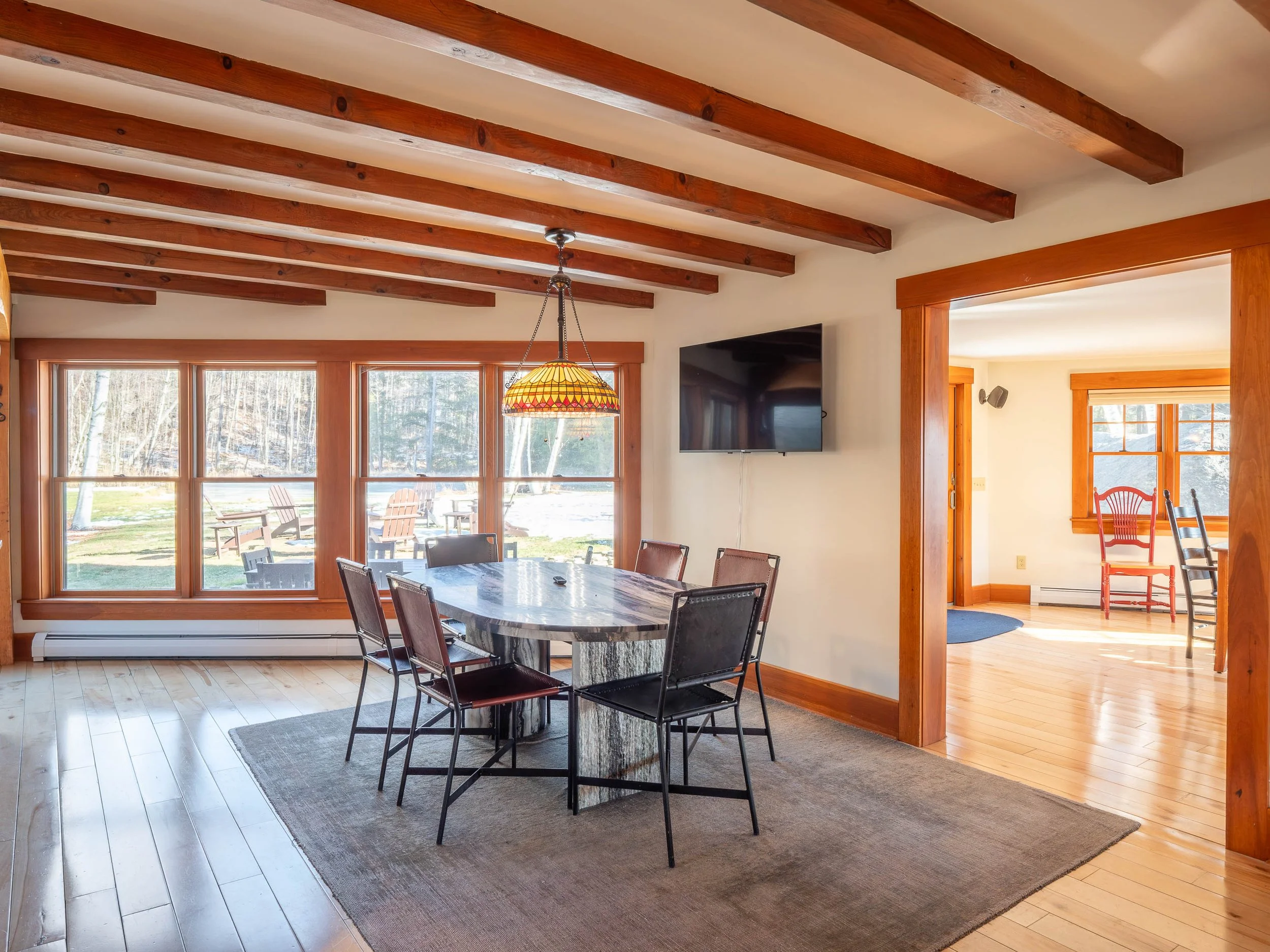 Dining area with a round table, six chairs, a Tiffany-style pendant lamp, wooden beams on the ceiling, large front windows, a wall-mounted TV, and hardwood floors, connecting to a brighter adjacent room.