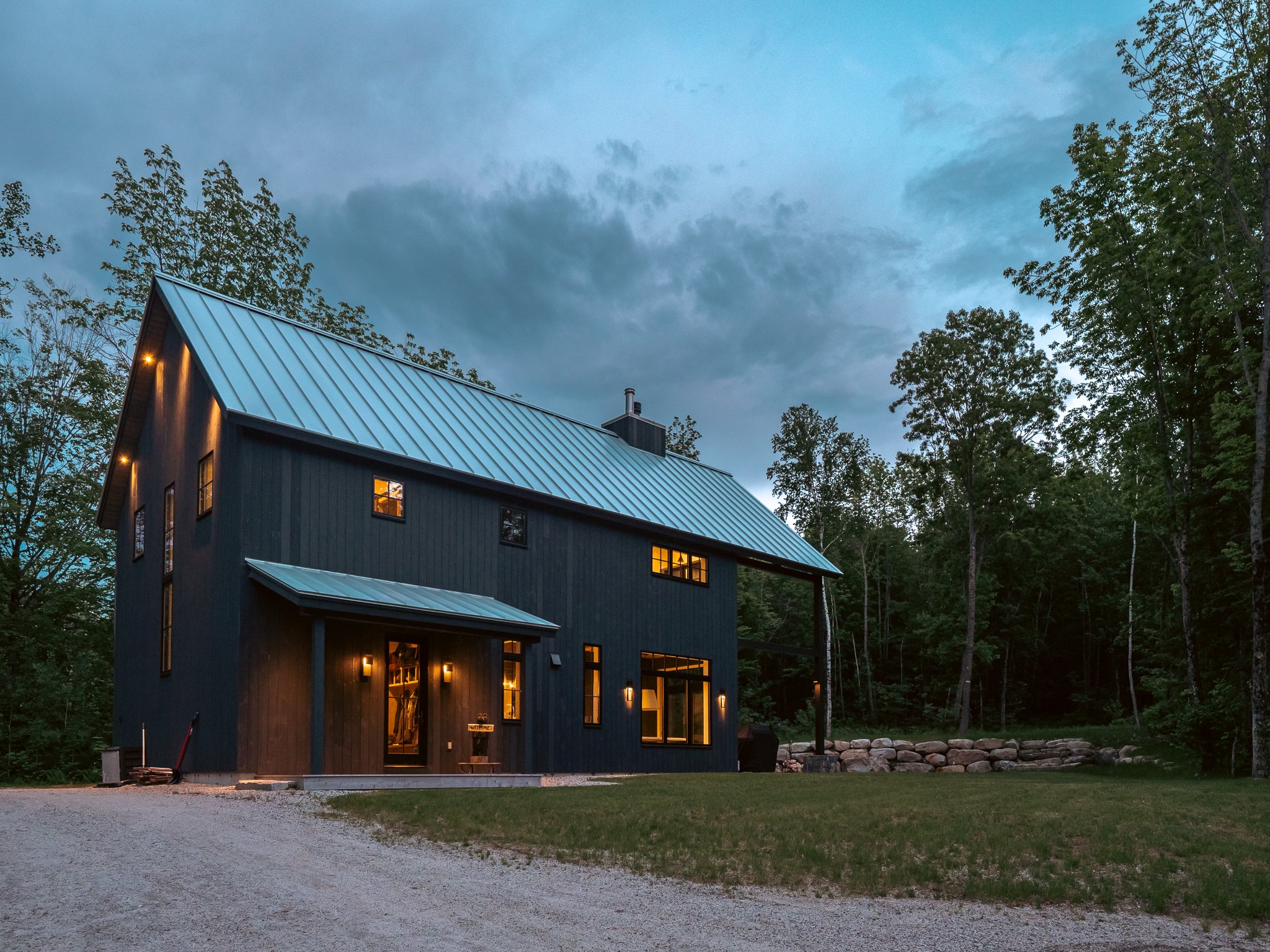 Modern barn-style house with metal roof and outdoor lighting, set against a wooded backdrop during twilight.