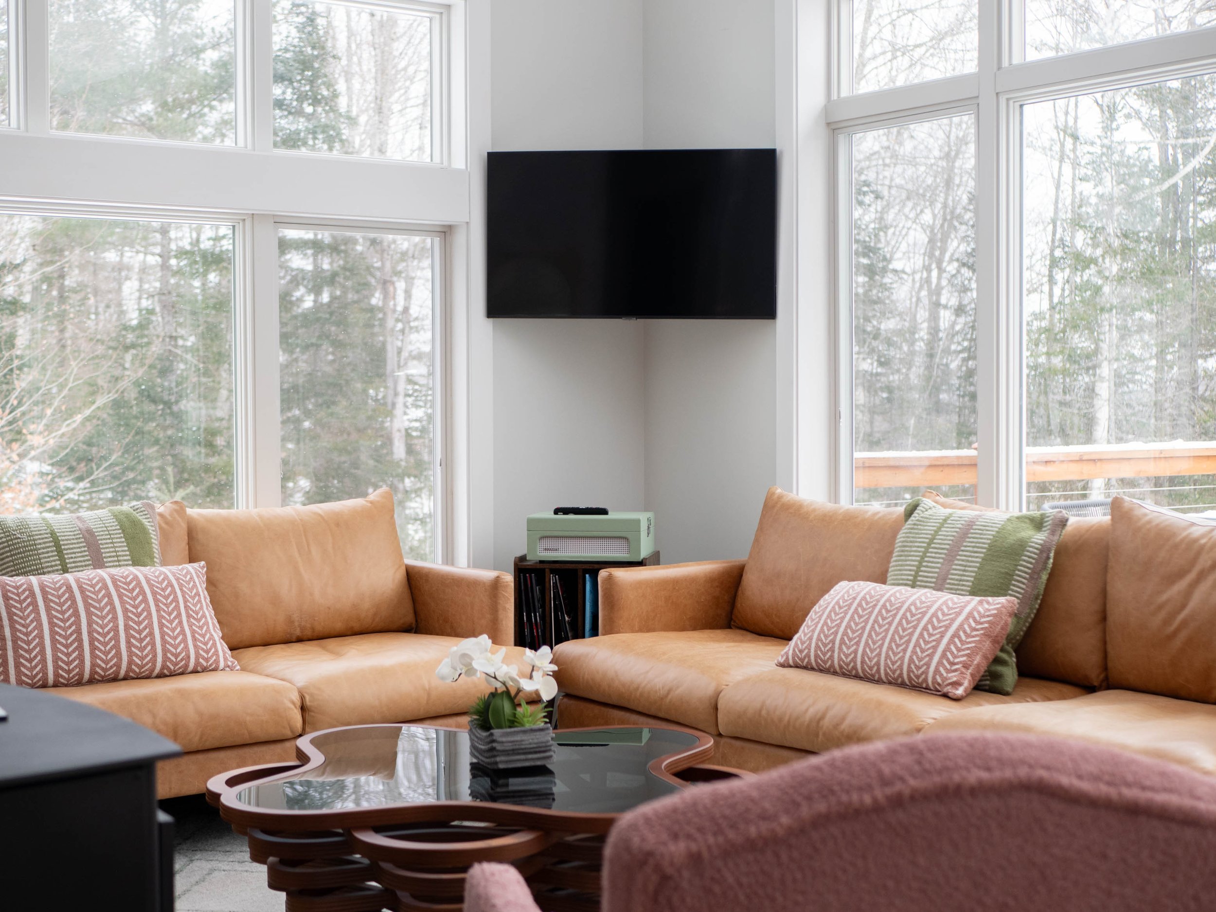 Living room with large windows, two tan sofas with patterned pillows, a black coffee table with a potted plant, and a wall-mounted TV.