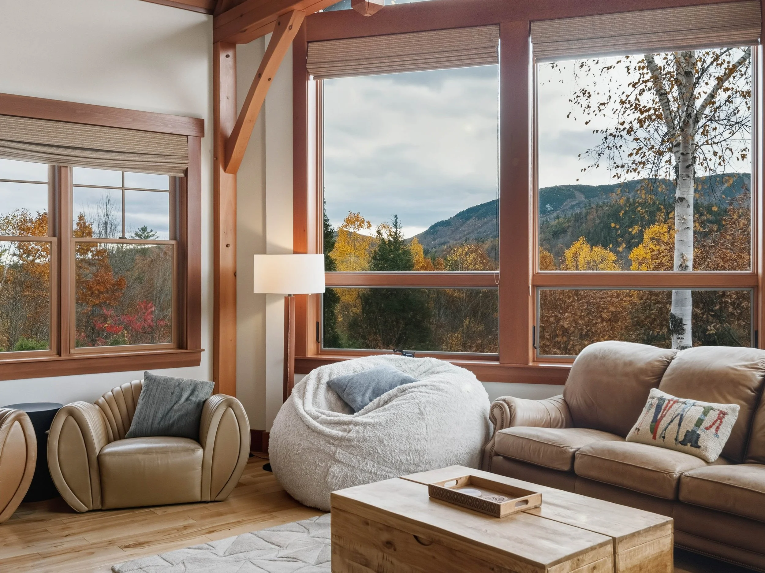 Living room with large windows showing an autumn landscape and mountains, beige leather sofa, cozy chair with throw pillow, large white bean bag, wooden coffee table, and tall lamp.
